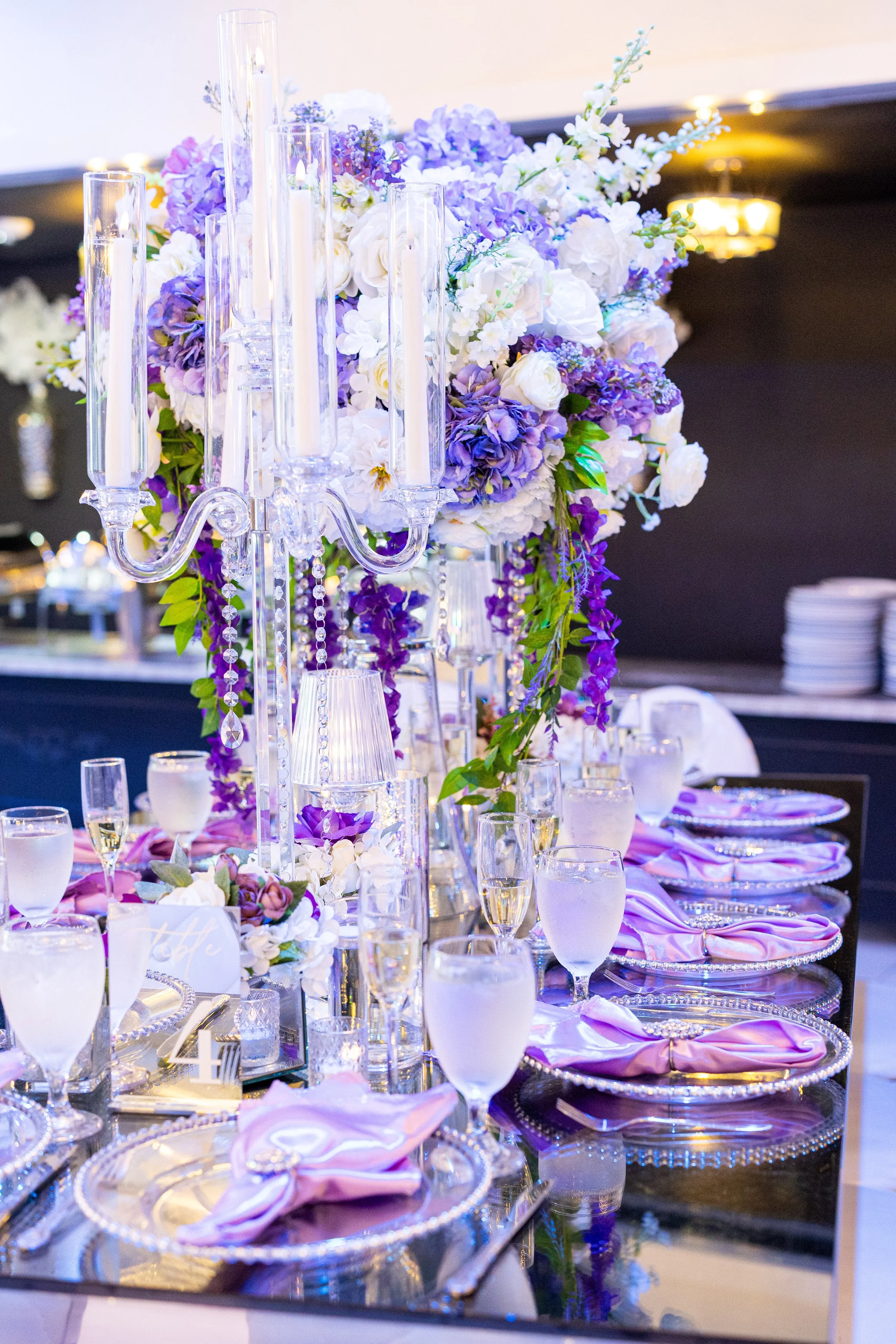 Elegant event table decoration with purple and white flowers, tall glass candle holders with lit candles, purple and pink napkins, and glassware on a reflective table surface.