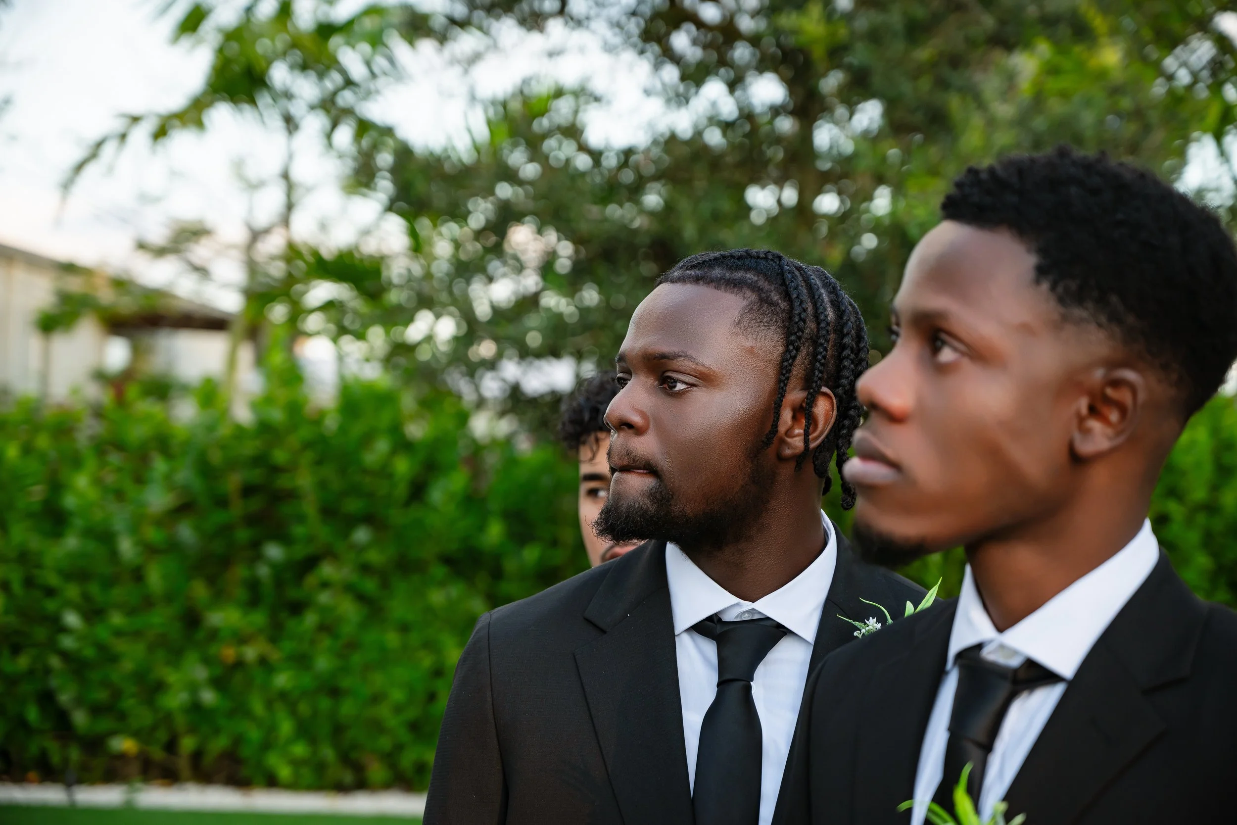 Three men wearing black suits and ties standing outside with greenery in the background, attending a formal event.