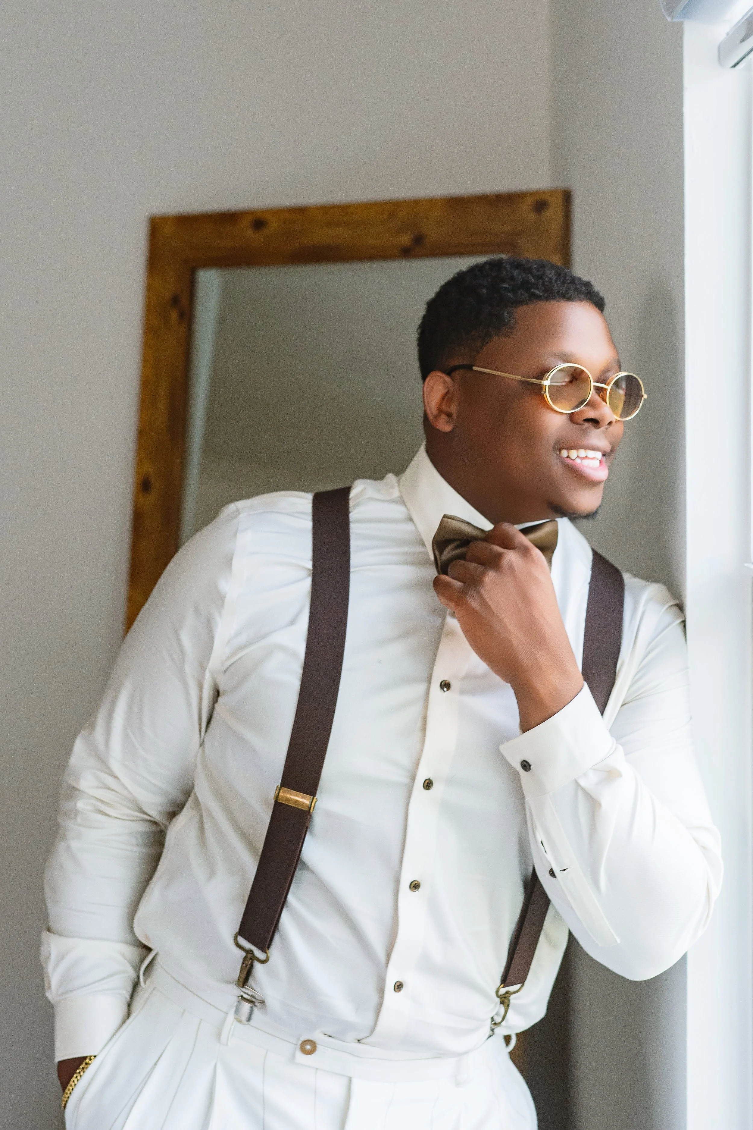 A stylish man dressed in a white shirt, suspenders, and a bow tie, looking out the window and smiling.