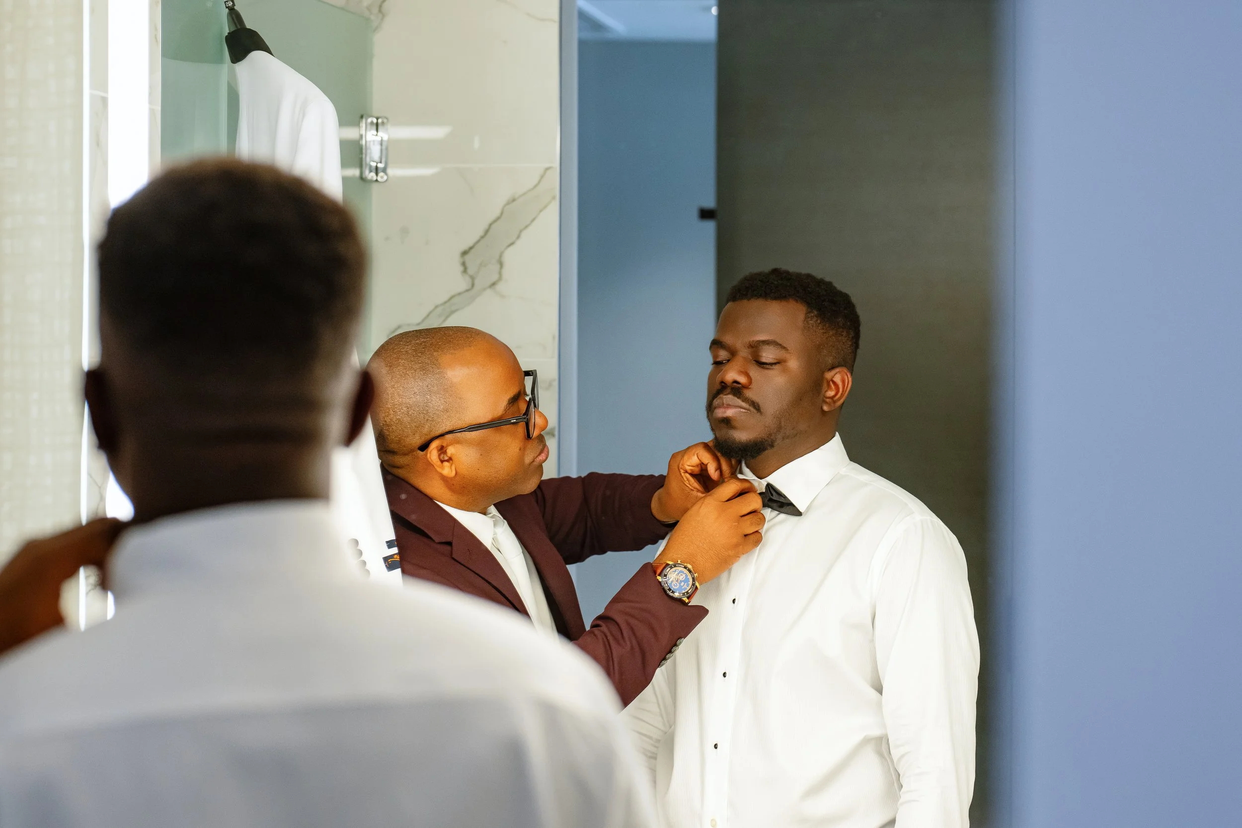A man in a white tuxedo coat is being assisted by another man in a maroon suit as he adjusts his bow tie, in a dressing room or bathroom with marble walls.