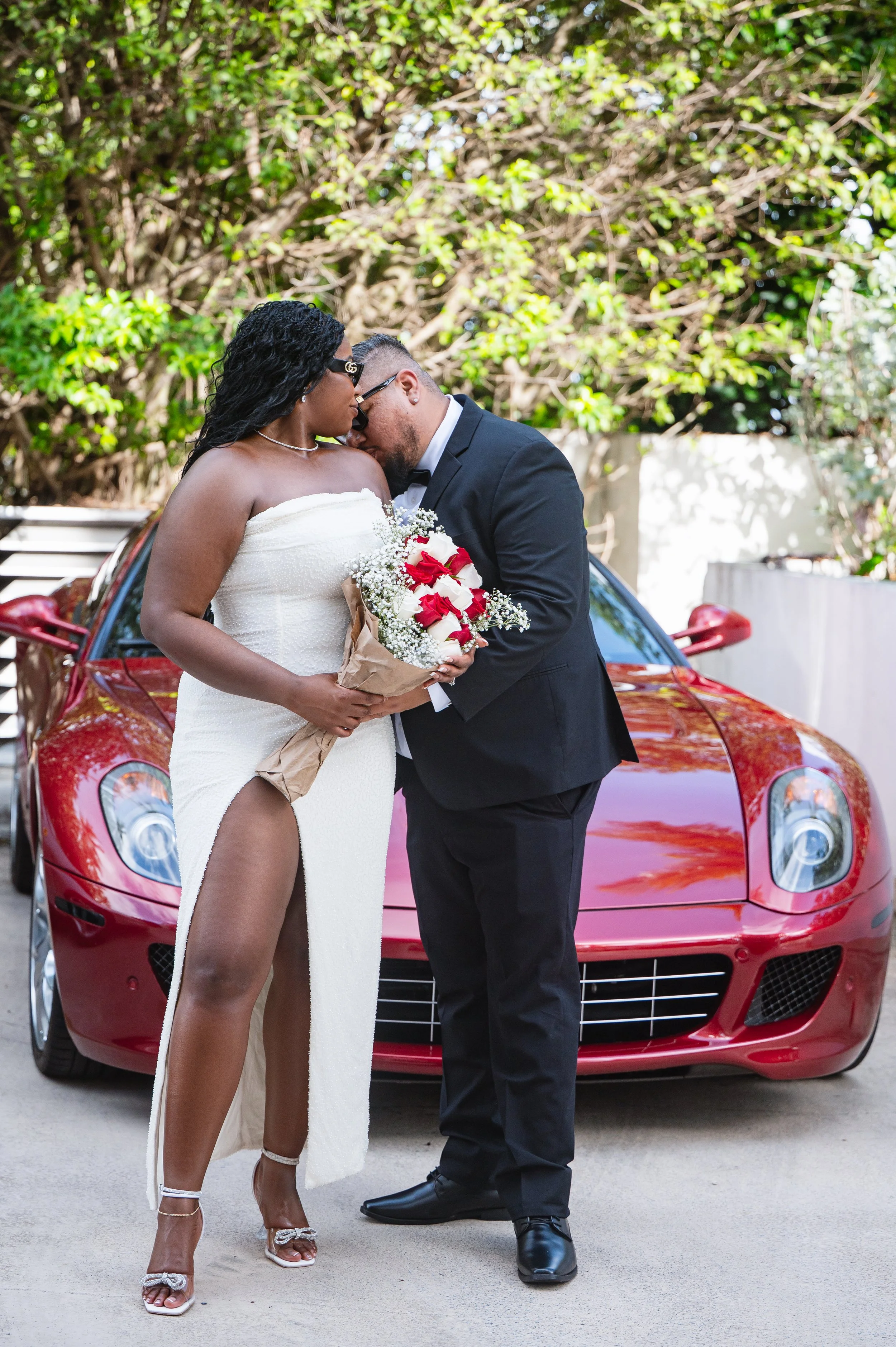 A couple dressed in wedding attire standing close together in front of a red sports car, with the woman holding a bouquet of white and red roses. They are outdoors with trees and greenery in the background.