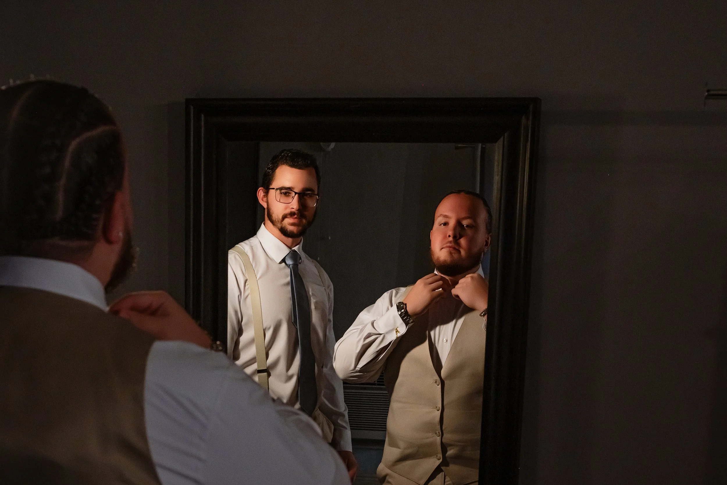 Two men looking at a mirror, one adjusting his collar and the other wearing glasses and suspenders, dressed in formal attire.