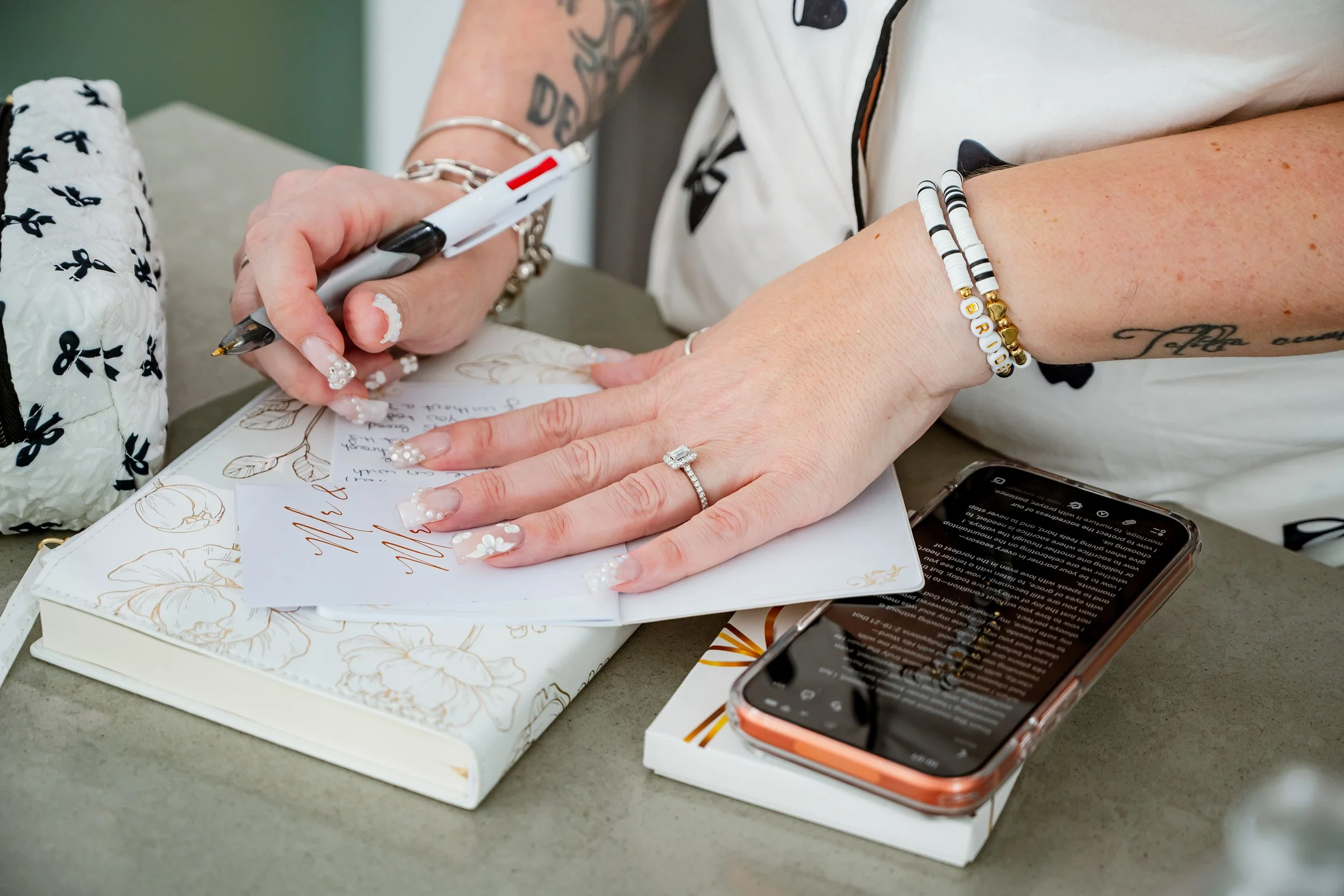A person with decorated nails and a ring on their finger is signing a wedding card with a black and white pen. They wear bracelets with beads and gold charms, and have a tattoo on their arm. There are wedding invitations, a smartphone, and a floral n