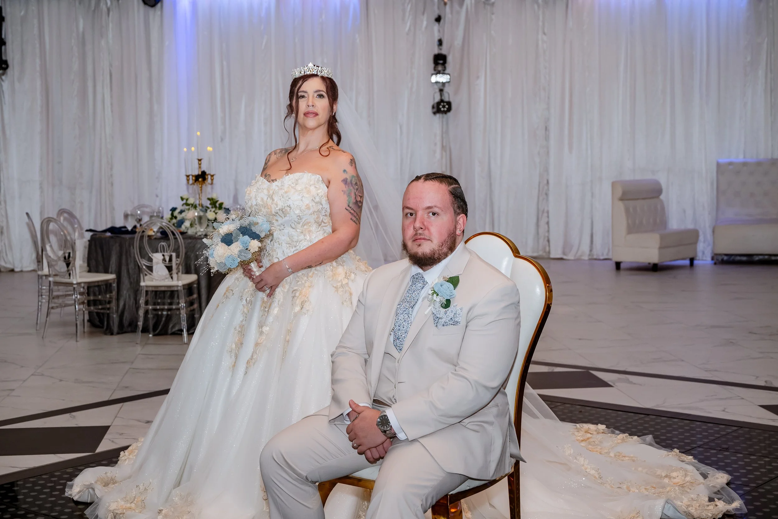 A newlywed bride standing beside a seated groom at their wedding reception. The bride wears a white wedding gown, crown, and holds a floral bouquet. The groom dons a white suit with a blue boutonniere. The background features a decorated event hall w