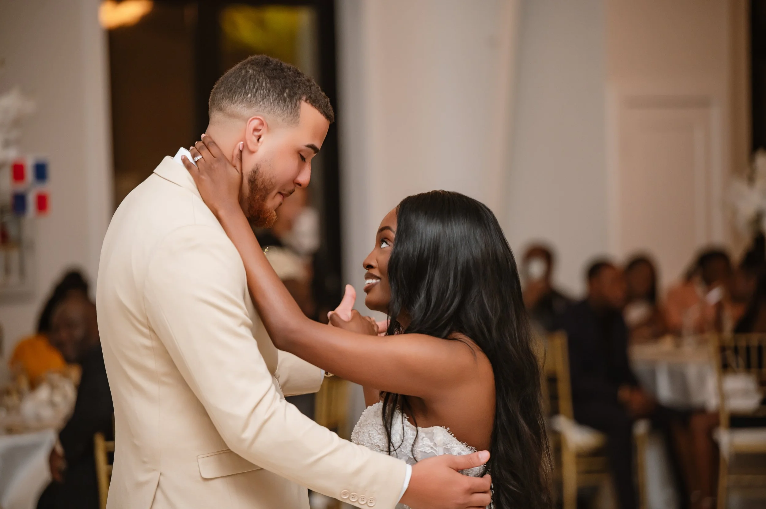 A couple, a man in a white suit and a woman in a white dress, dancing closely at a wedding reception, with guests in the background.