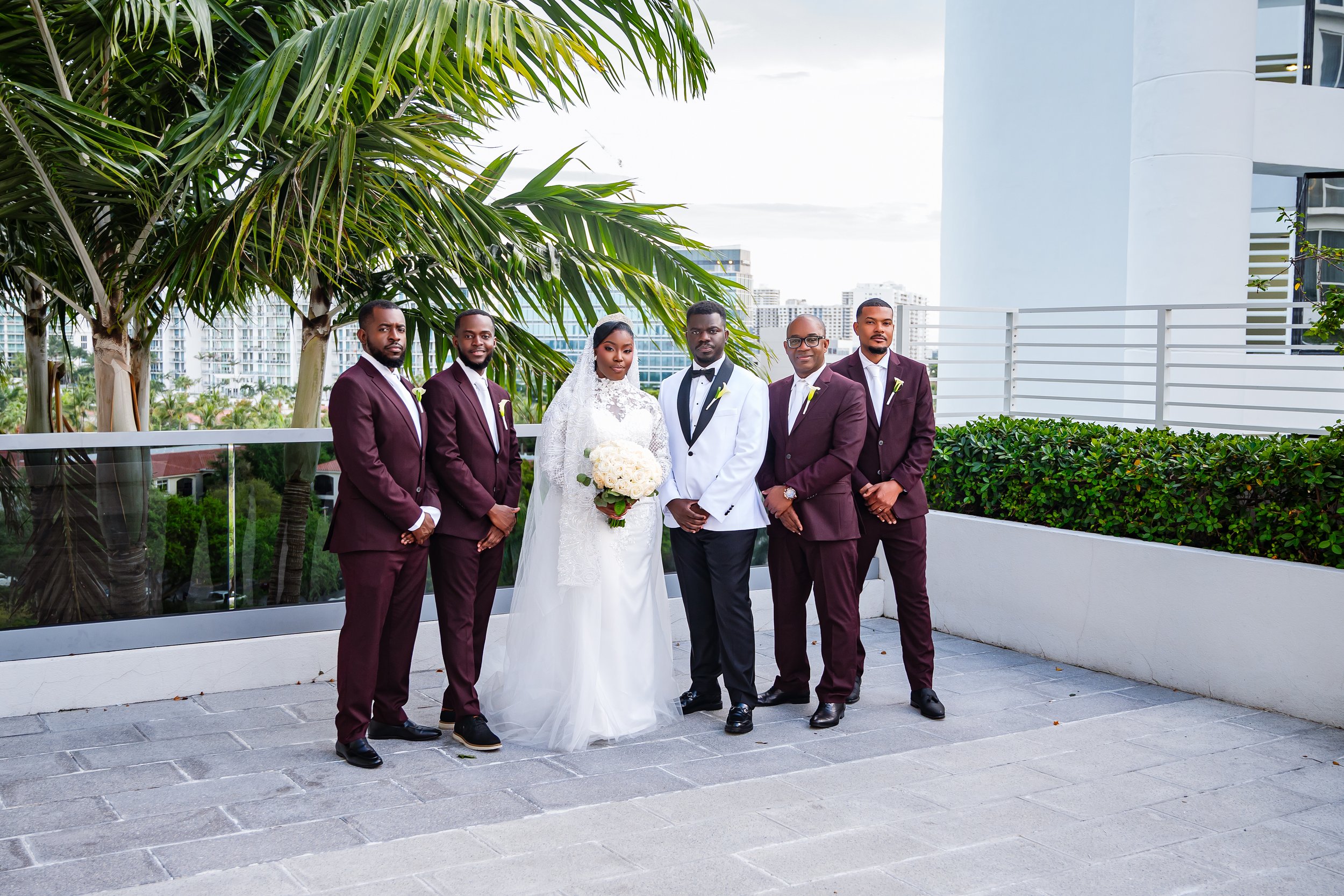 A bride in a white lace dress holding a bouquet standing with five men in matching maroon suits on a rooftop terrace with greenery and city buildings in the background.