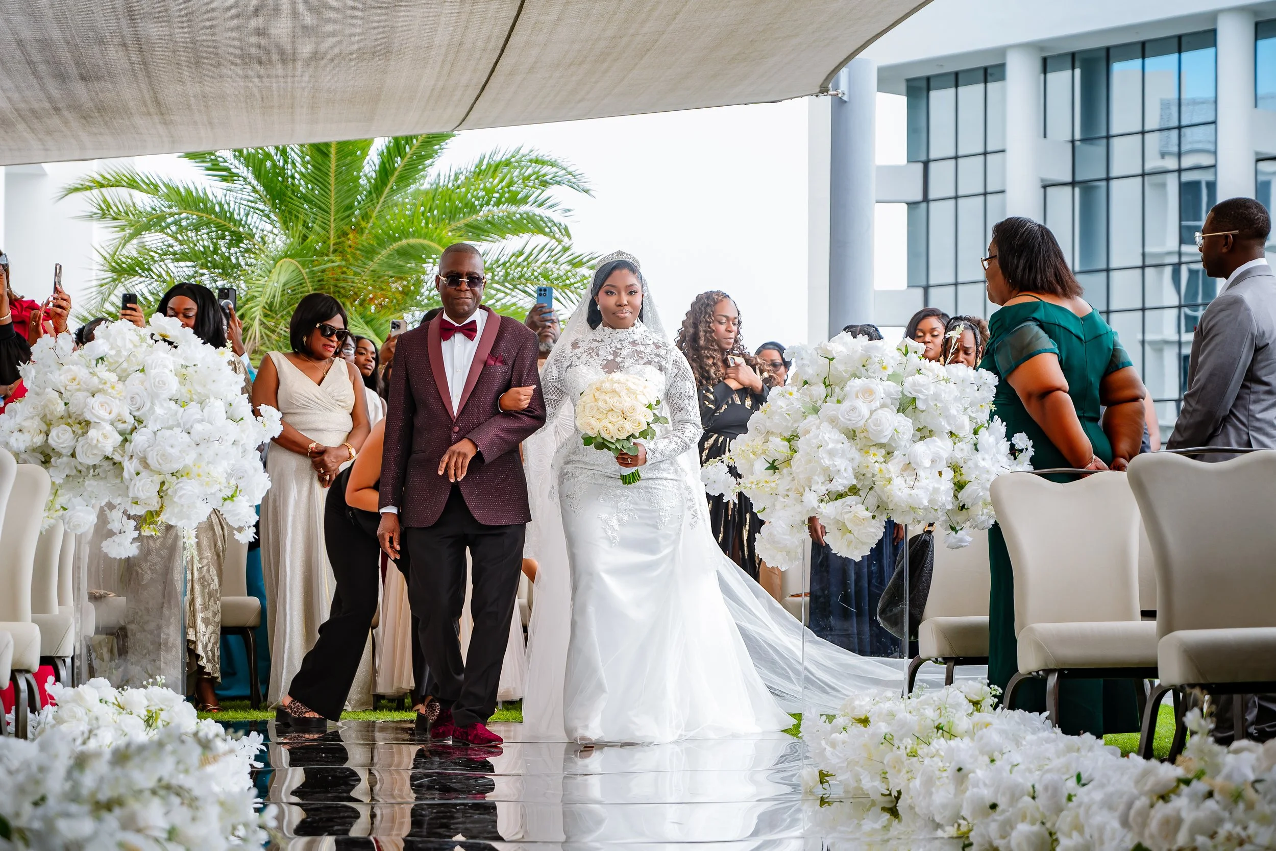 A bride in a white wedding dress with veil holding a bouquet of white roses, walking down the aisle accompanied by a man in a burgundy suit and sunglasses at an outdoor wedding with guests taking photos and a modern building in the background.