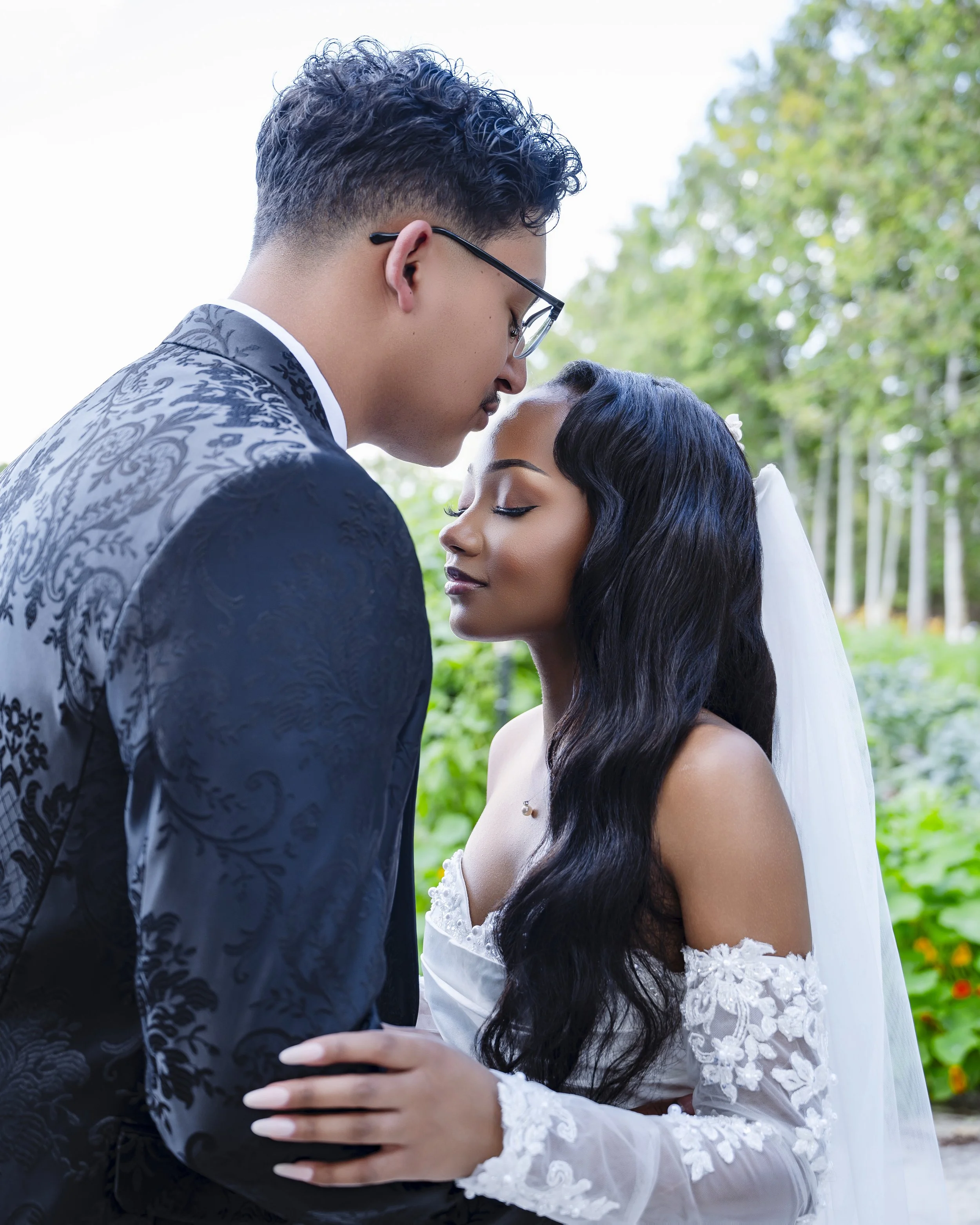 A couple in wedding attire standing close with foreheads touching outdoors, greenery in the background.
