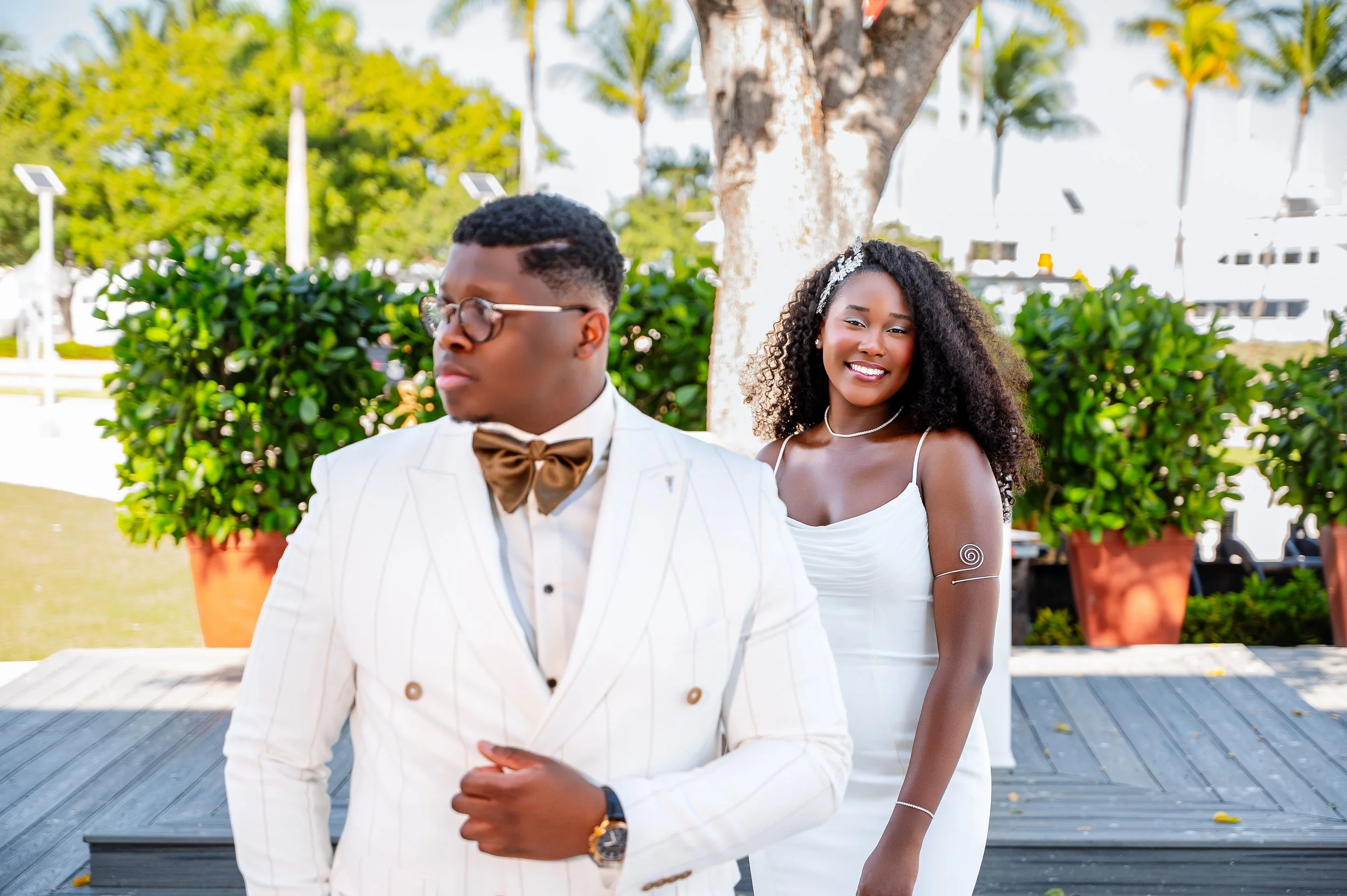 A man in a white suit with a brown bow tie and glasses stands in the foreground, looking away. A woman in a white dress with curly hair and a headband smiles behind him, standing outdoors near a tree and large potted plants.