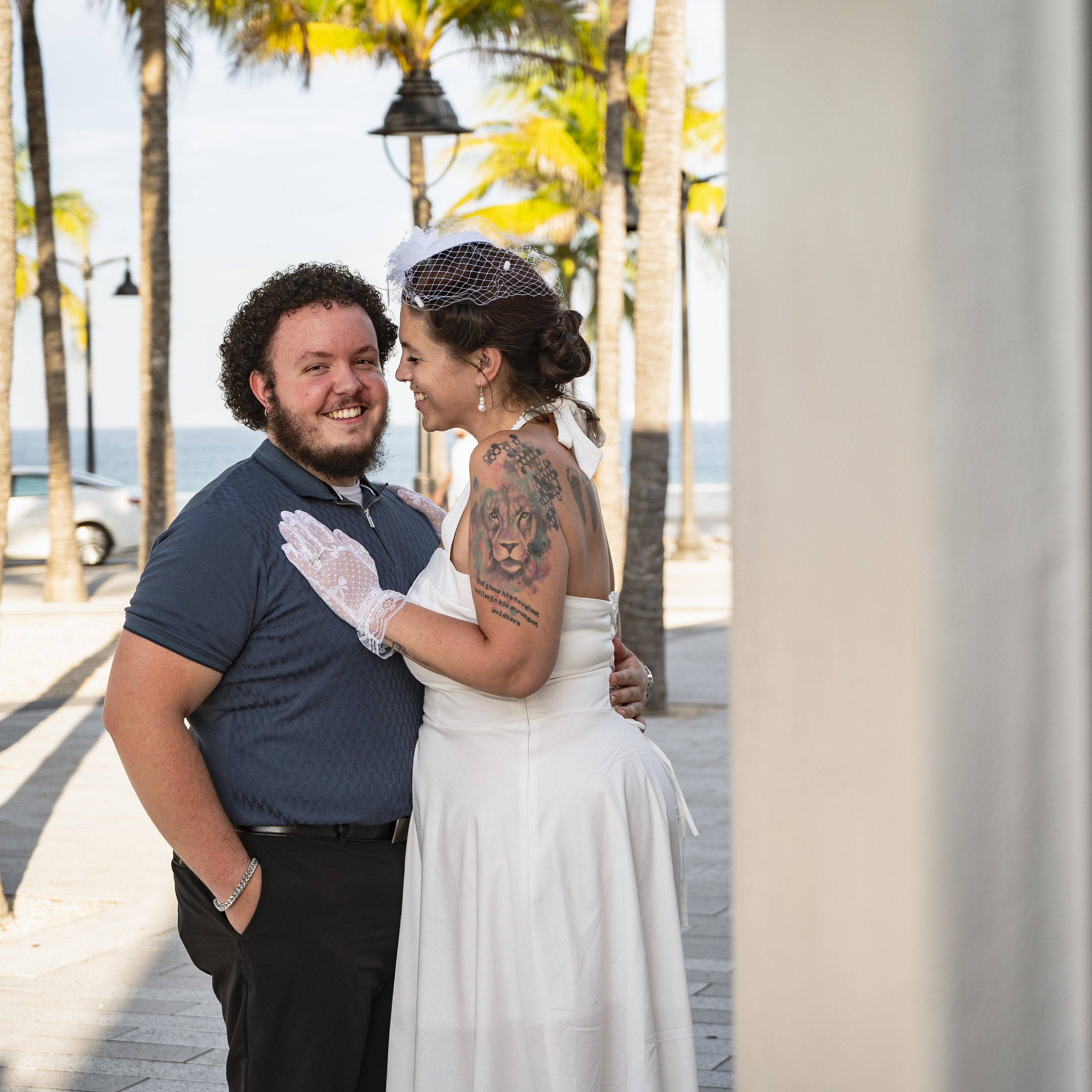 A happy couple, dressed for a wedding, standing outdoors near palm trees with the ocean in the background. The bride has tattoos, wears a white dress, veil, and gloves, and is leaning into the groom, who is smiling.