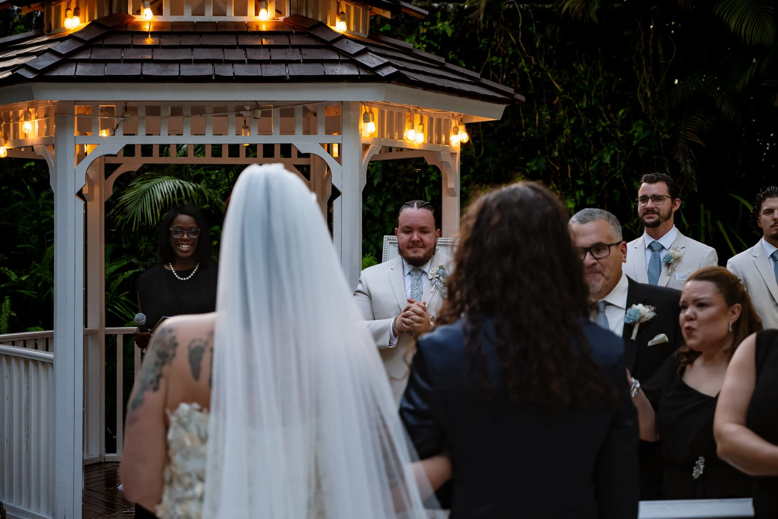 A wedding ceremony outdoors with a bride dressed in white and guests standing in front of a decorated gazebo surrounded by greenery.