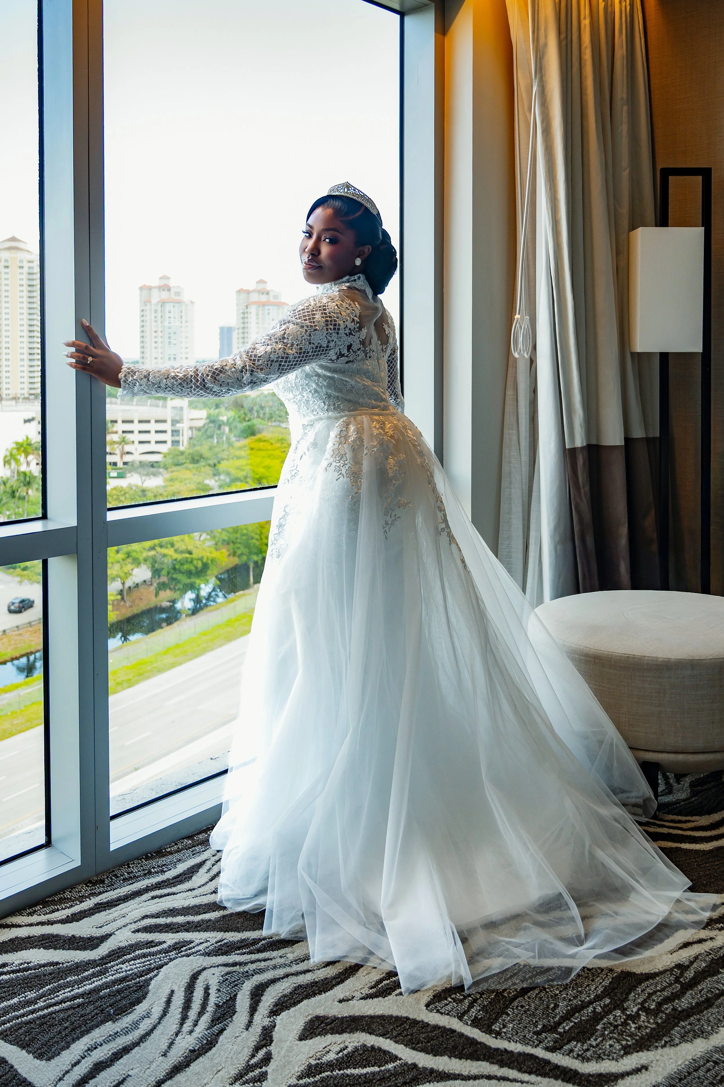 A woman in a wedding dress standing near a large window with city buildings outside, holding the window frame and looking at the camera.