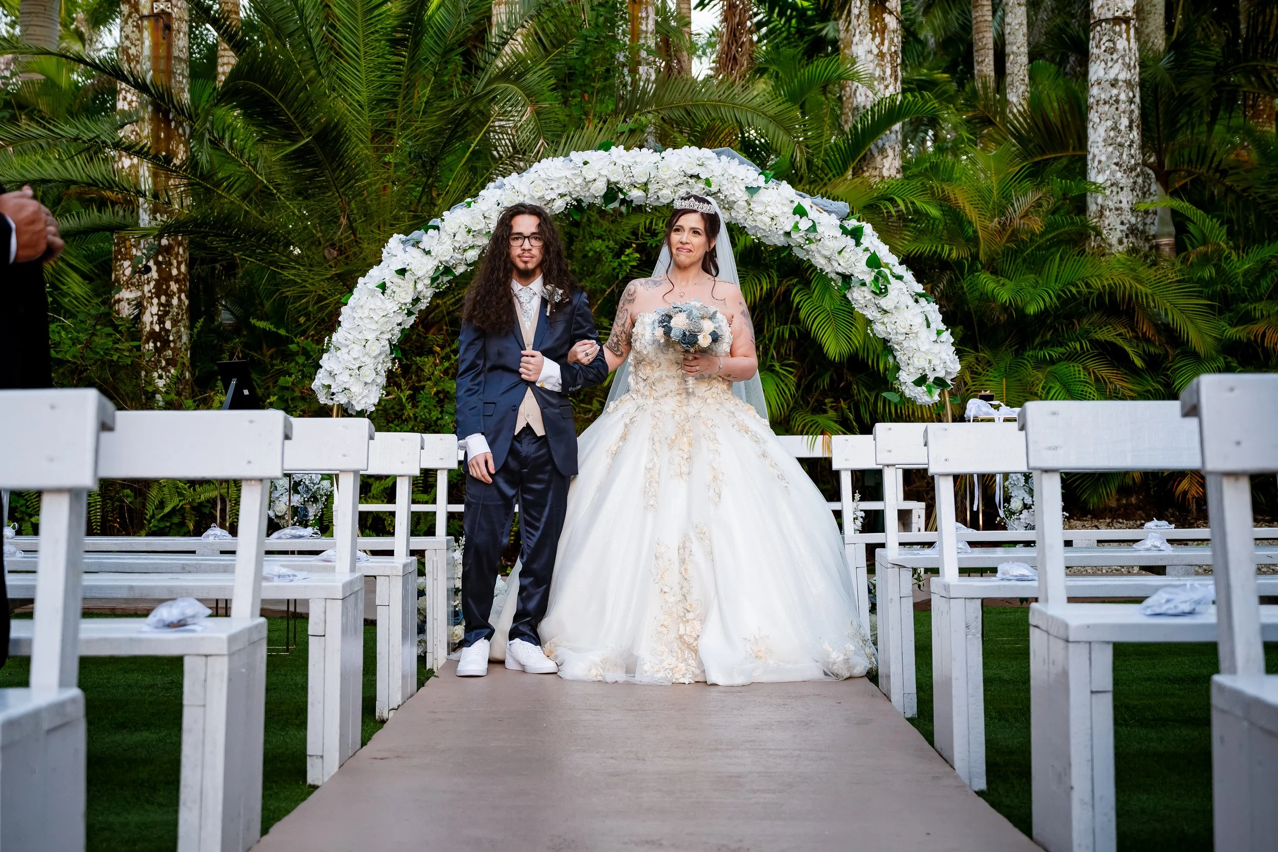 A bride and groom standing together at an outdoor wedding ceremony, with the bride holding a bouquet and wearing a gown, and the groom in a navy suit and white sneakers, in front of an arch decorated with white flowers and surrounded by lush green fo