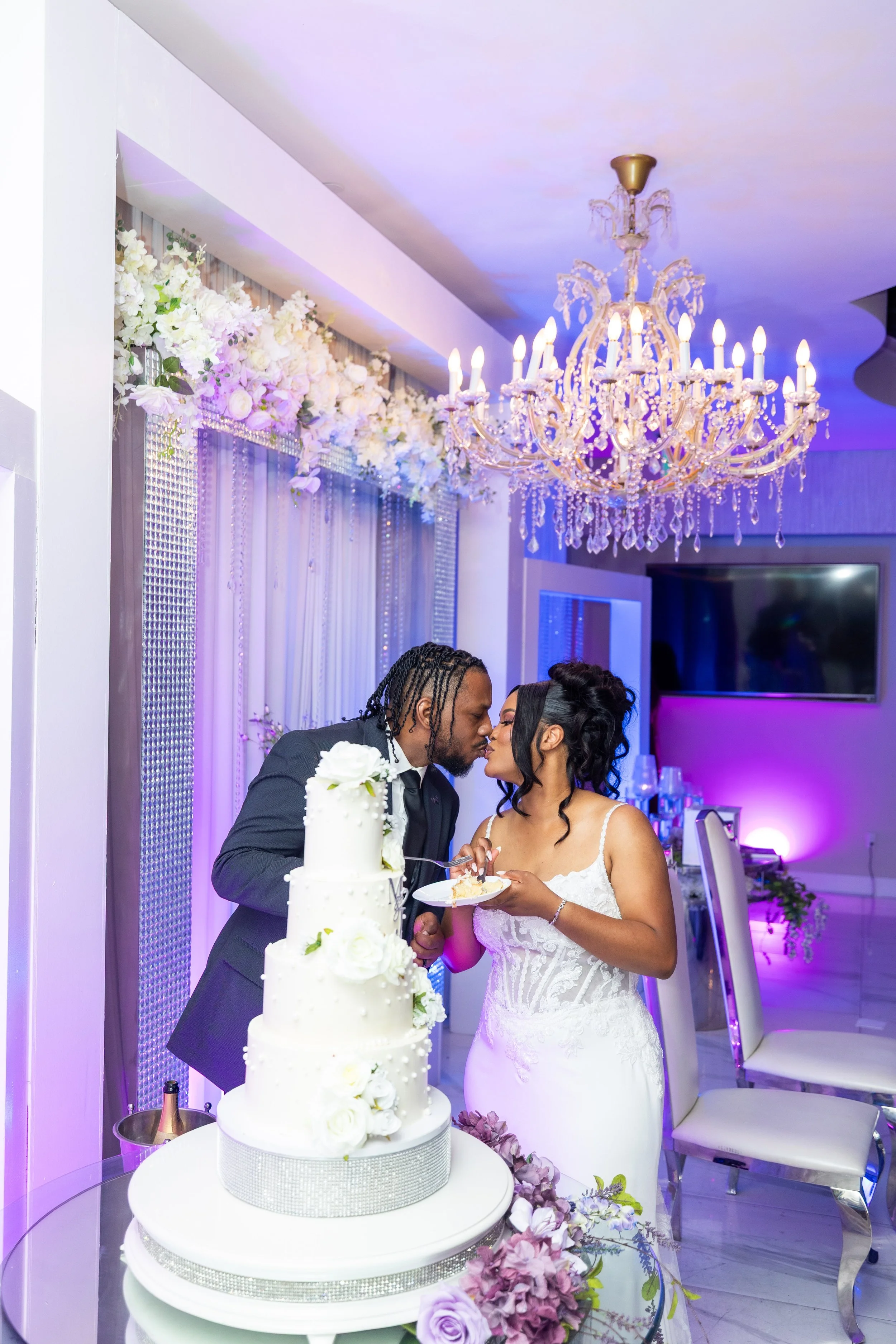 A bride and groom are standing near a multi-tiered white wedding cake with floral decorations, sharing a moment at their wedding reception. The room is decorated with a chandelier, floral arrangements, and ambient purple lighting.