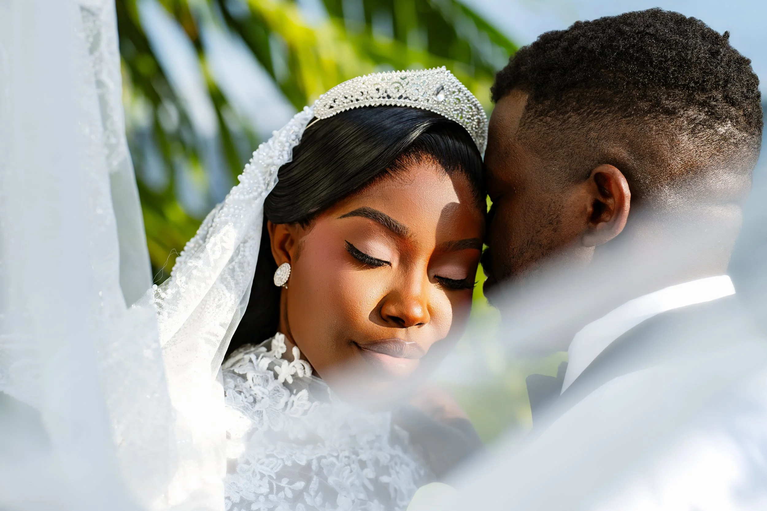 Close-up of a bride and groom with their foreheads touching, eyes closed, during a wedding ceremony outdoors. The bride wears a white lace dress and a tiara with pearls, and the groom wears a white shirt. Green foliage and a blurred background are vi