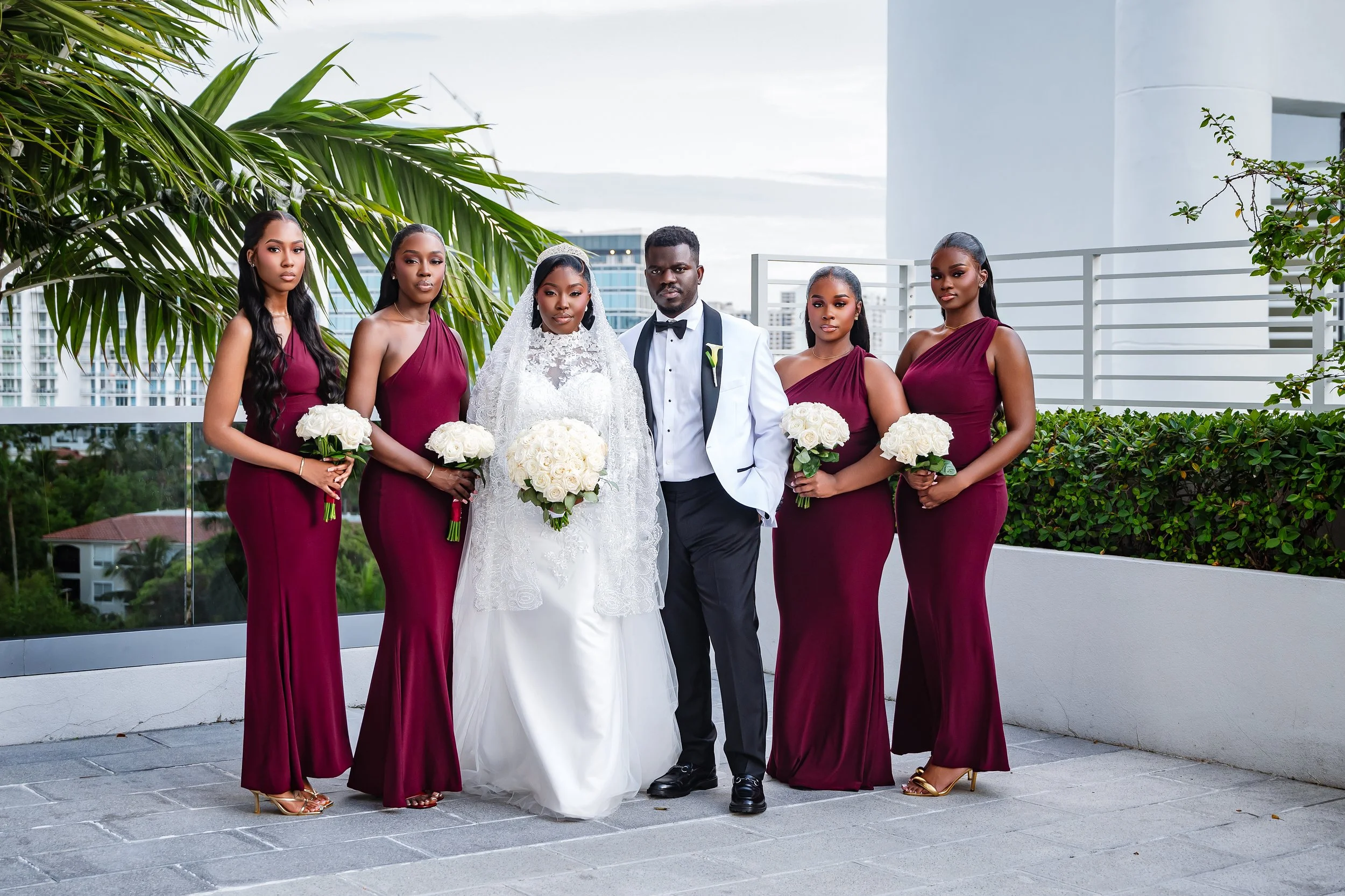Group of six people, including a bride in a wedding dress holding a bouquet of white roses, and five women in maroon dresses holding similar bouquets, standing on a terrace with modern buildings and greenery in the background.