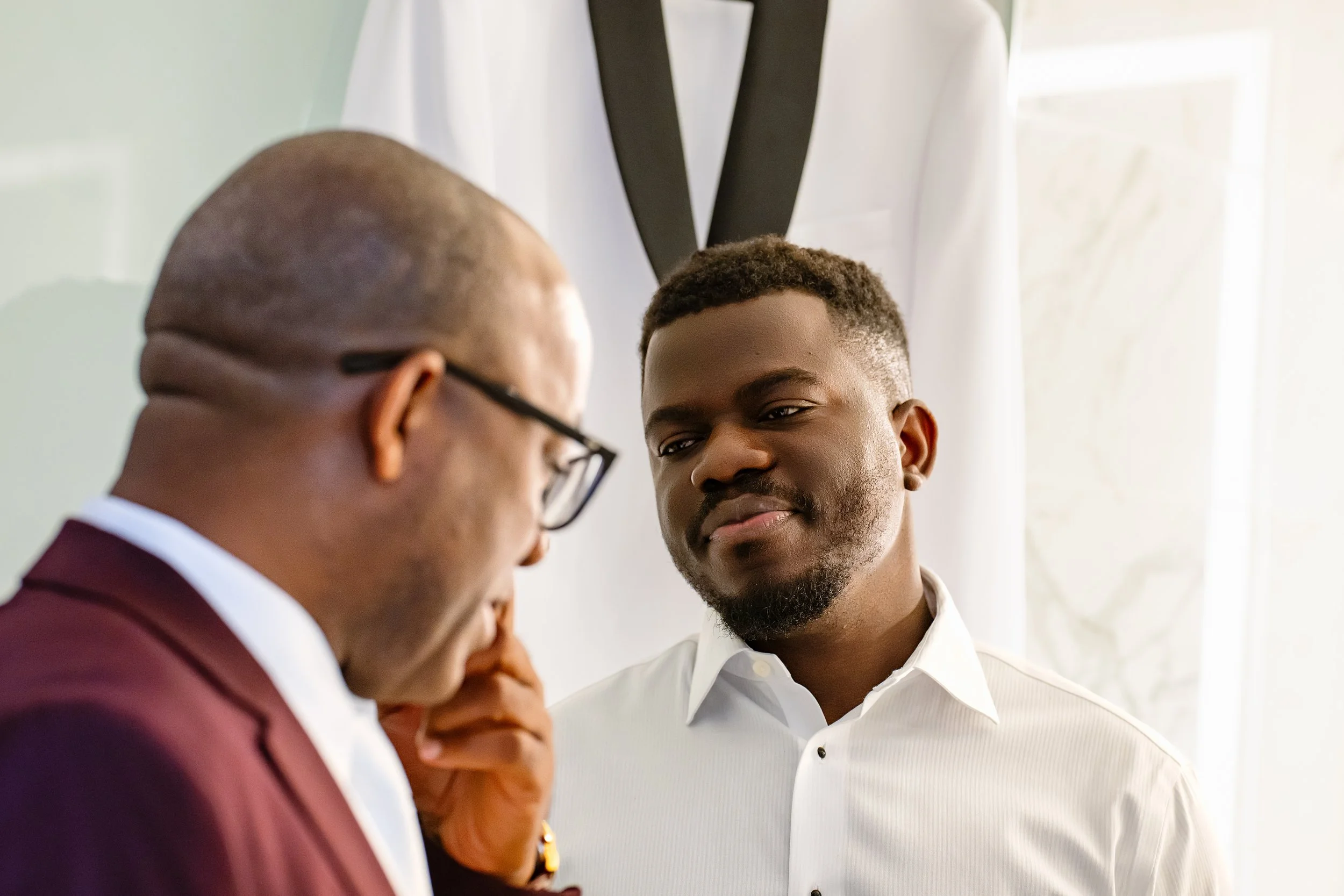 A young man with short curly hair and a beard looking down at an older man with glasses, who is partially visible. They are in a room with natural light and wearing formal clothing.