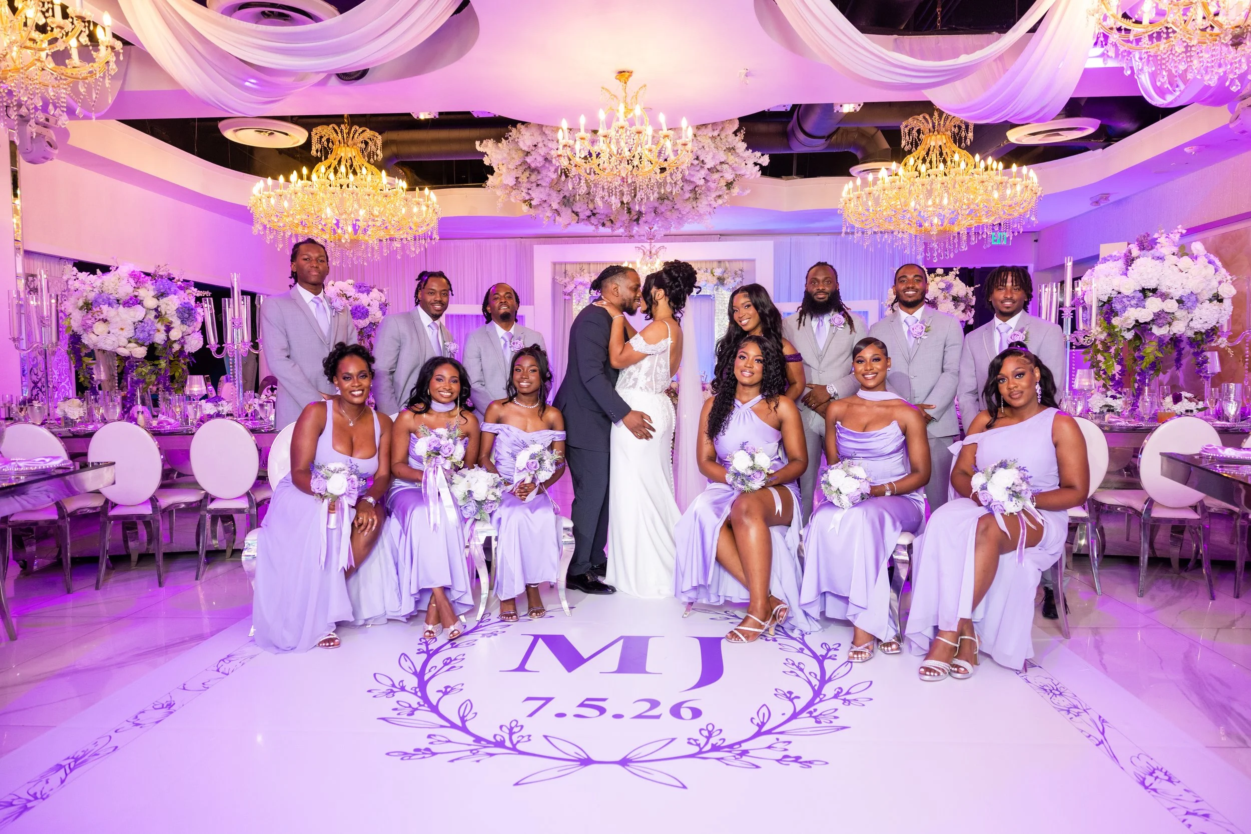 Group of people celebrating a wedding in a decorated venue with chandeliers and purple lighting. The bride and groom stand in the center, surrounded by bridesmaids in lavender dresses and groomsmen in gray suits. The floor has a monogram with the ini