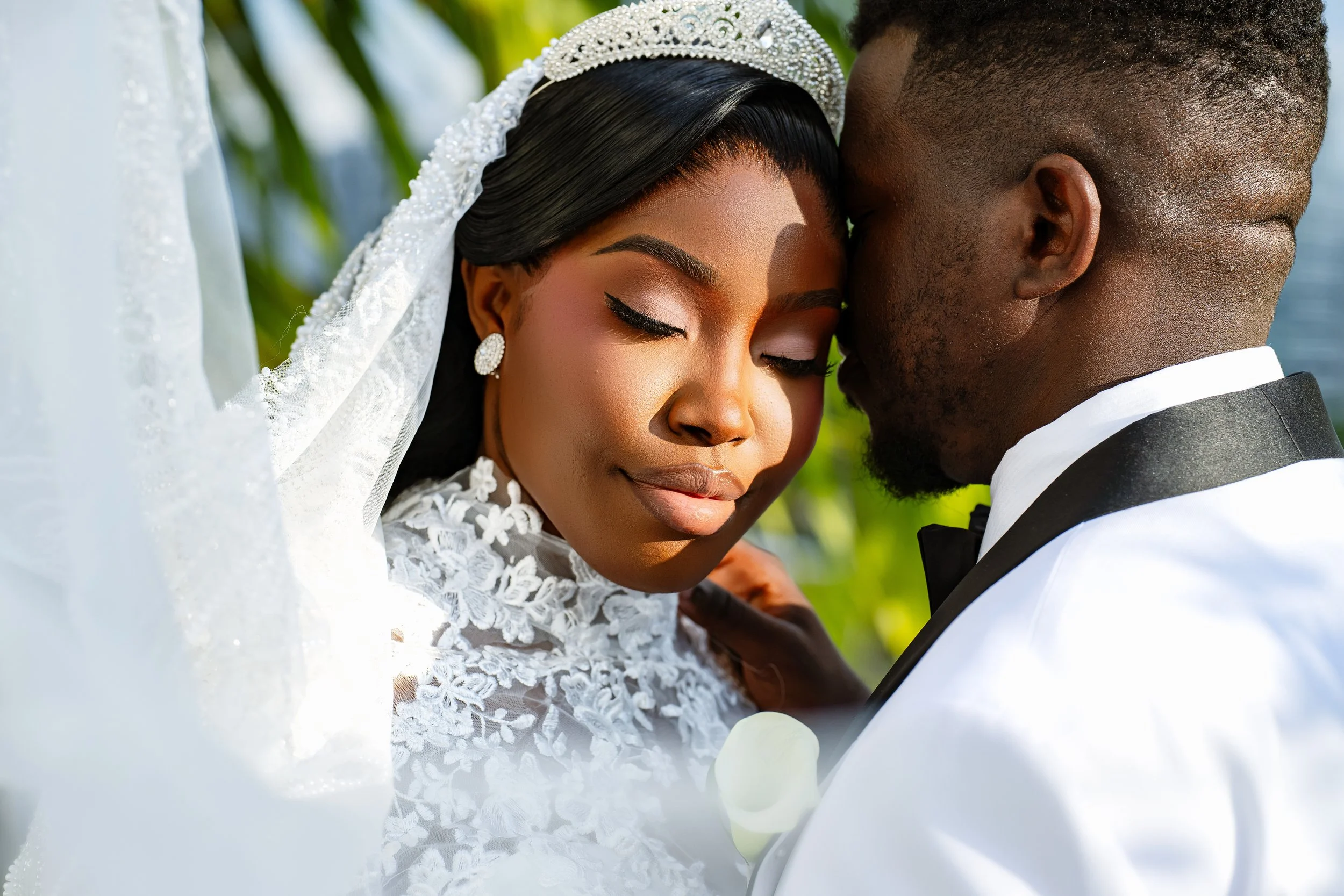A bride and groom close together, with eyes closed, during a wedding ceremony outdoors, with greenery in the background.
