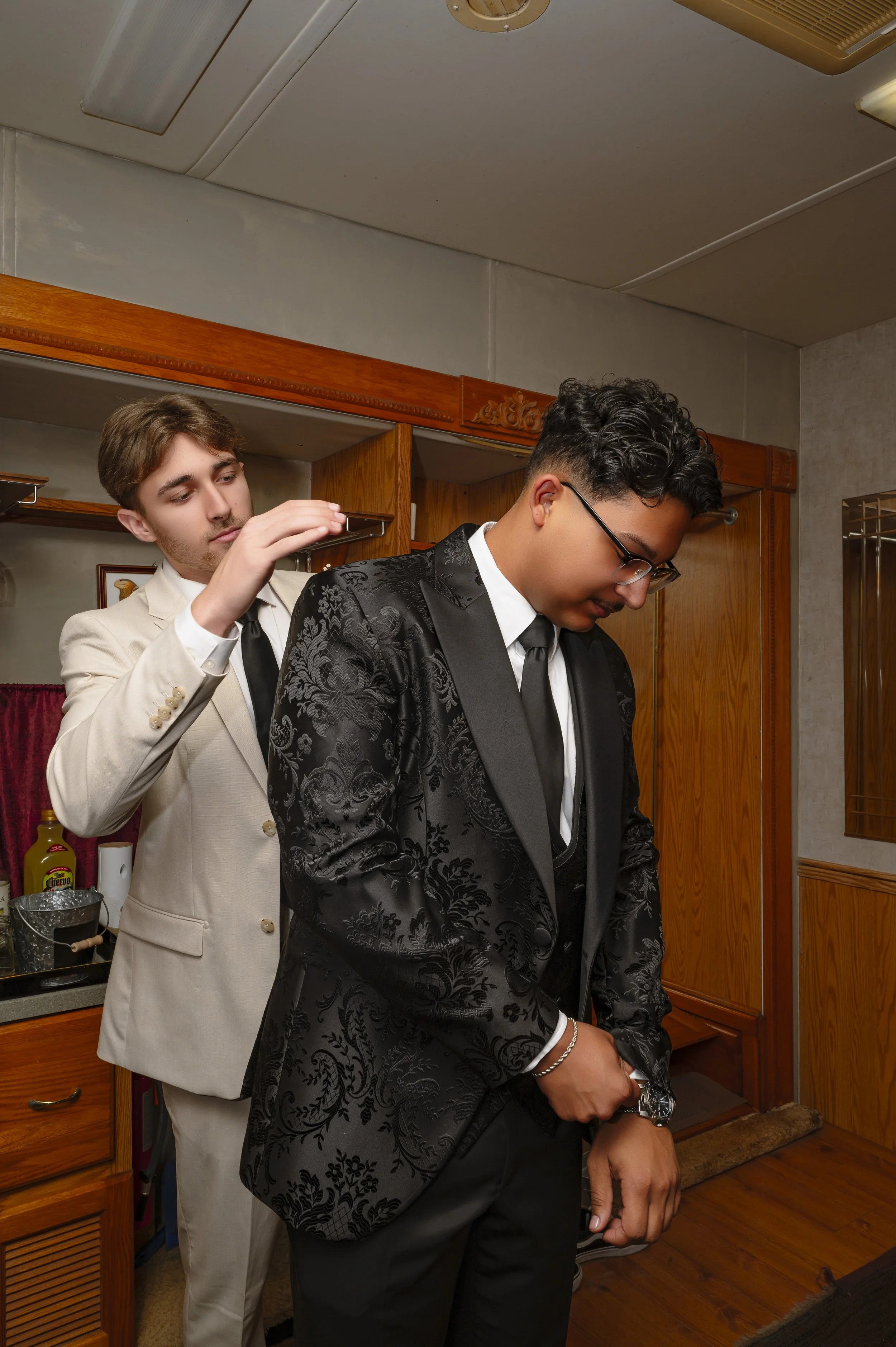 Two men, dressed in formal suits, are preparing for a formal event. One man, with glasses and a black floral-patterned suit, is standing with his head slightly bowed. The other man, in a beige suit, is adjusting the bow tie of the first man.