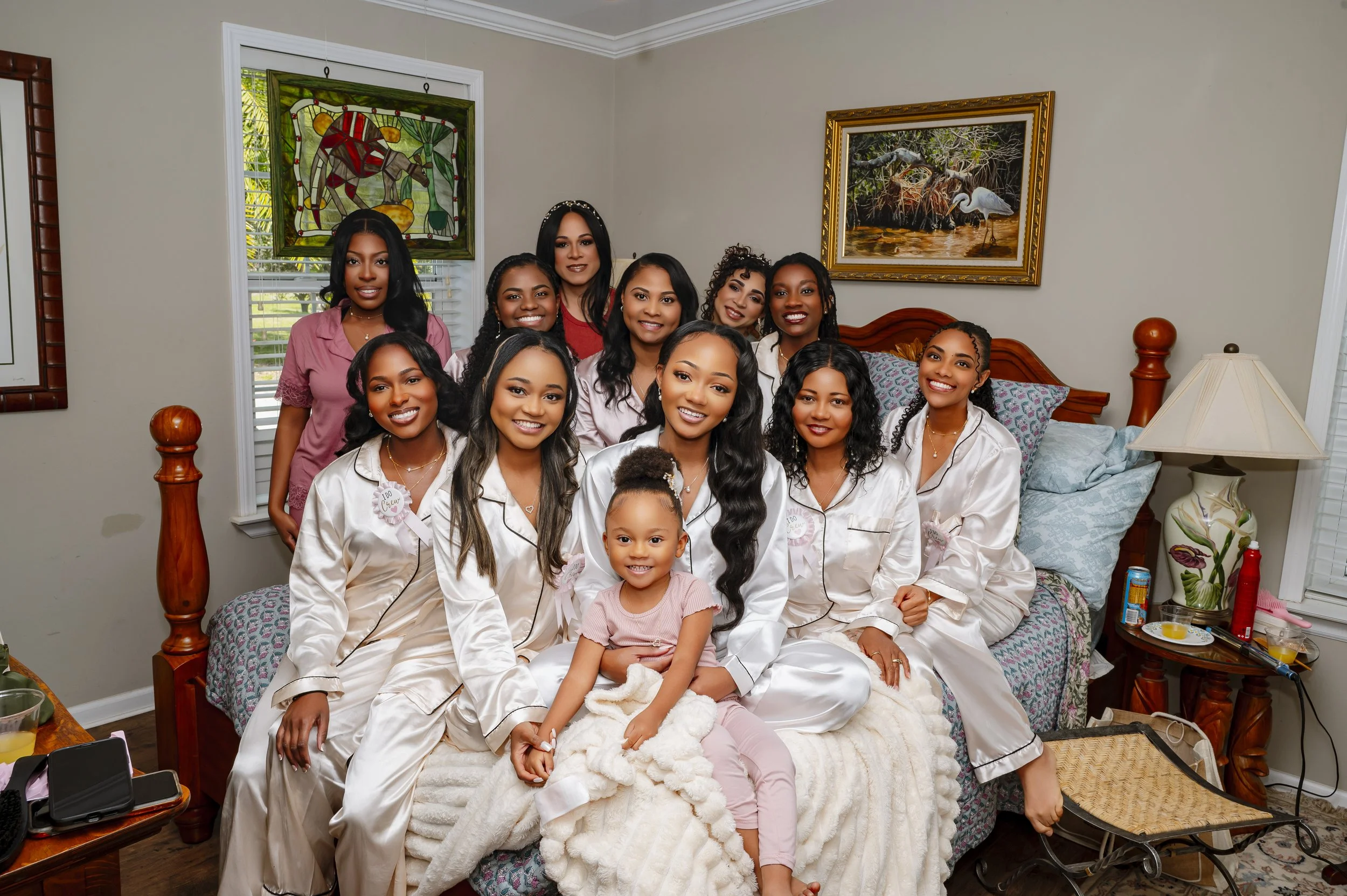 A group of women and a young girl sitting and standing on a bed in a bedroom, posing for a photo.