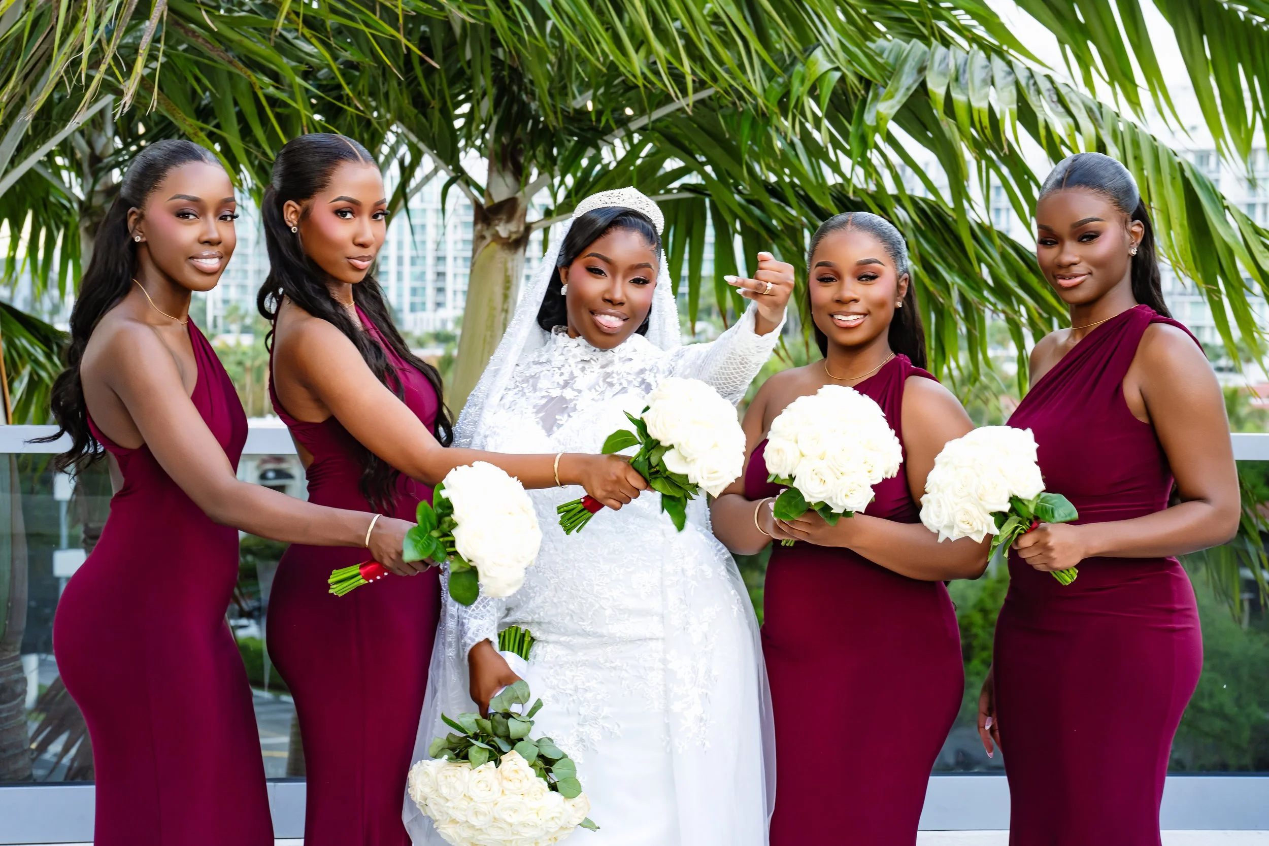 Bride in a white wedding dress with veil and tiara holding a bouquet of white roses, standing between five bridesmaids wearing matching burgundy dresses and holding white rose bouquets, outdoors under green trees, with city buildings in the backgroun