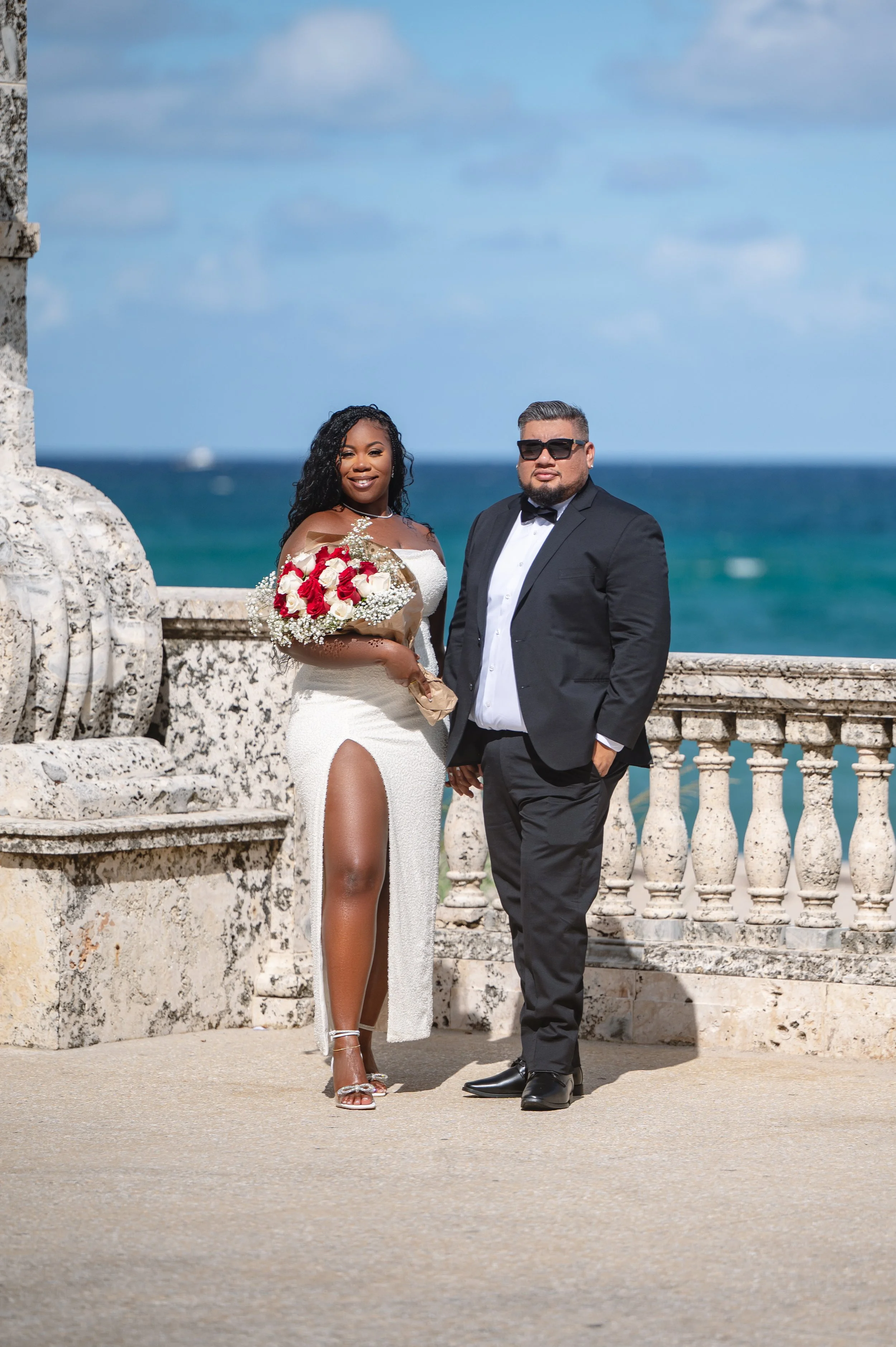 A bride and groom on a seaside terrace, with a blue ocean and sky in the background. The bride is wearing a white off-shoulder gown with a high slit, holding a bouquet of red and white roses. The groom is dressed in a black tuxedo with sunglasses, st