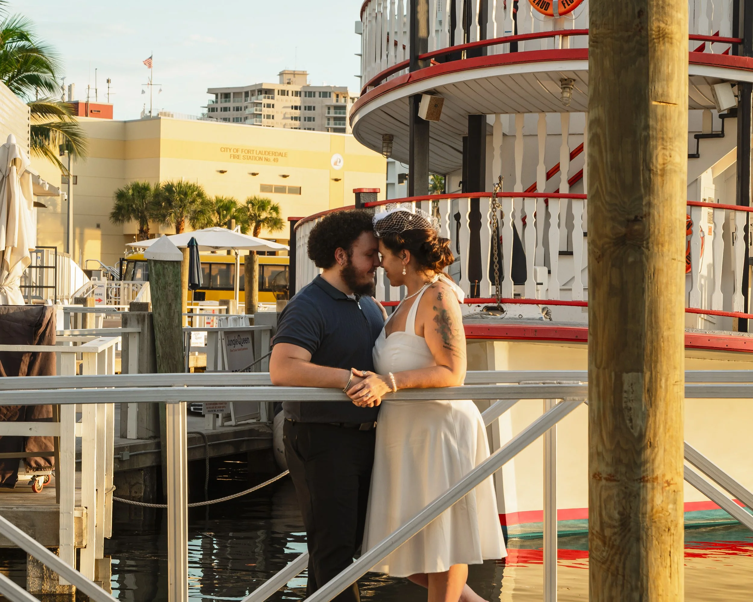 A couple stands close with foreheads touching on a dock near a boat, surrounded by urban buildings and palm trees during sunset.
