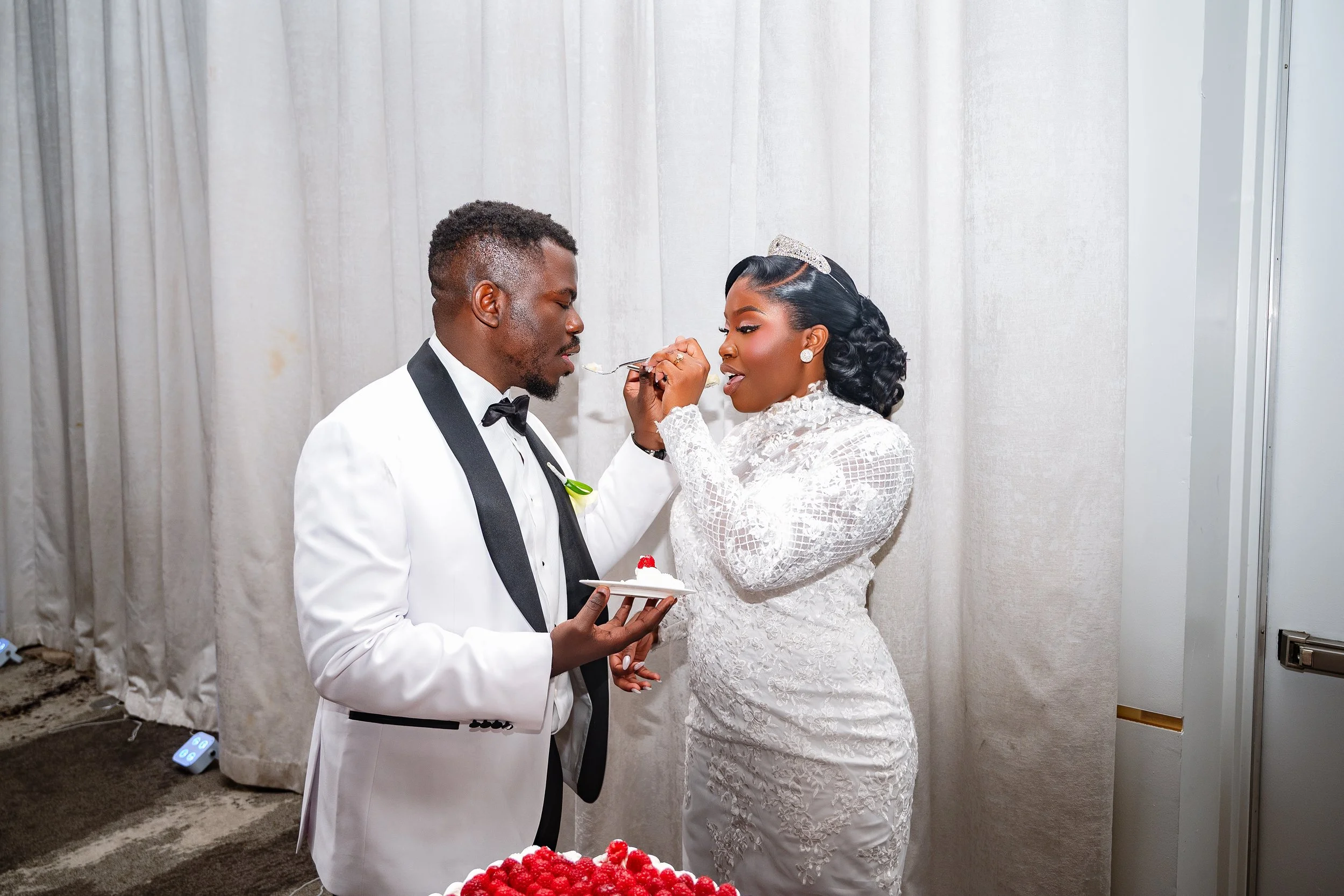 A bride and groom in wedding attire sharing a cake cutting, with the bride feeding the groom a piece of cake using a fork, and the groom holding a plate with some cake and a fork, against a backdrop of white curtains.