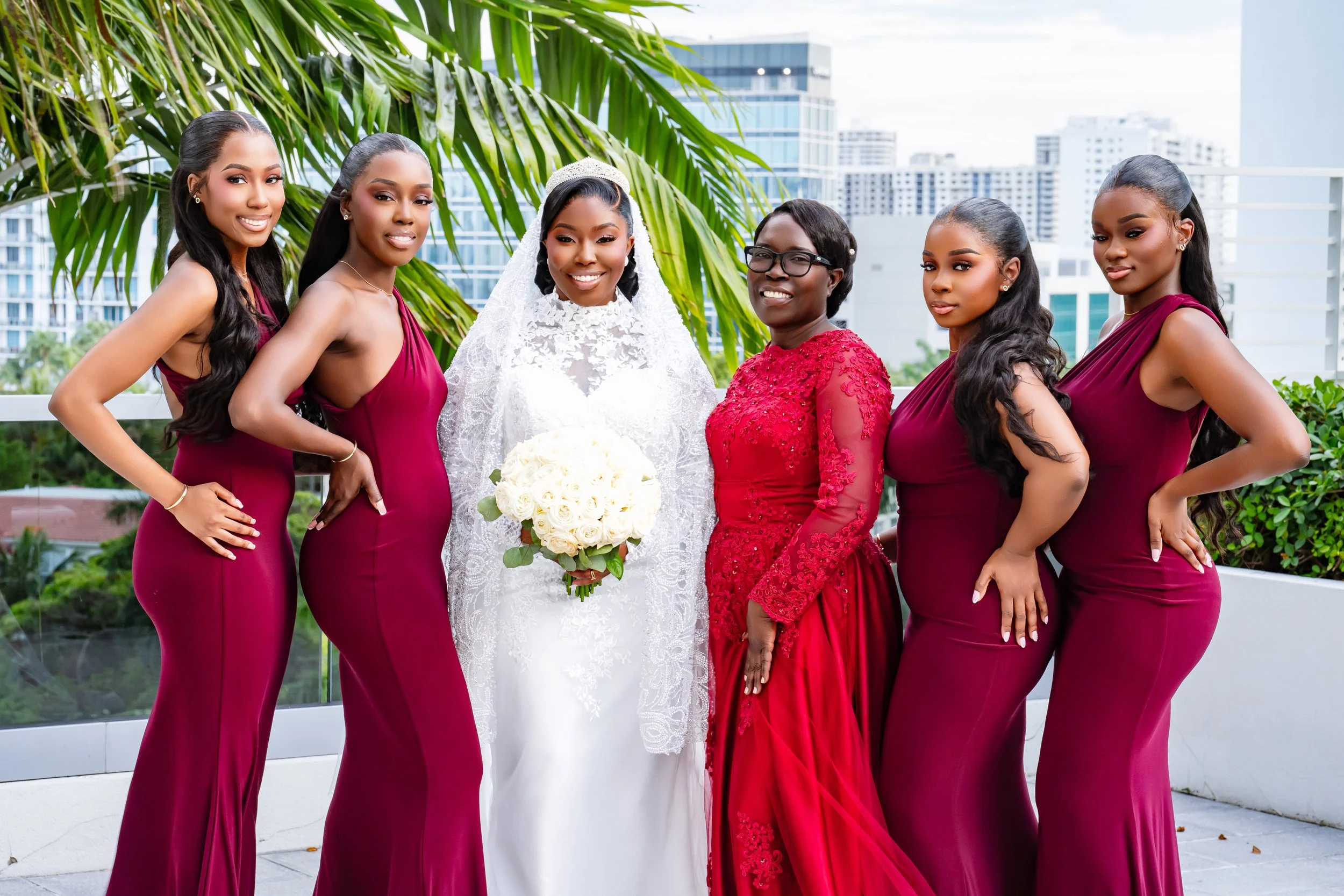 A bride in a white lace wedding dress holding a bouquet of white roses and five bridesmaids in matching maroon dresses posing on a balcony with city buildings in the background.