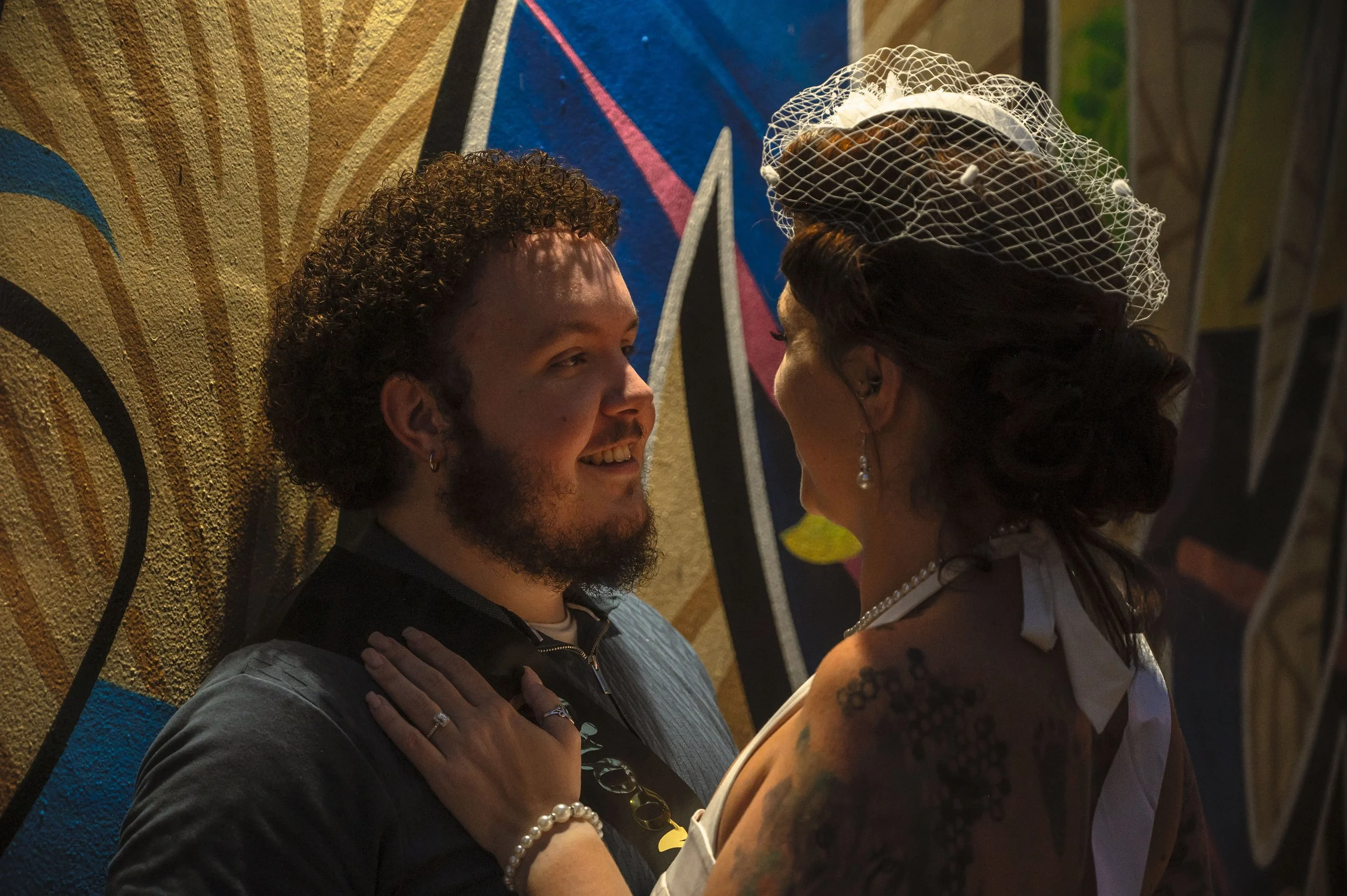 A couple faces each other closely, smiling, with one person having a tattooed arm and wearing a vintage hat, jewelry, and a wedding dress, standing in front of a colorful graffiti wall.