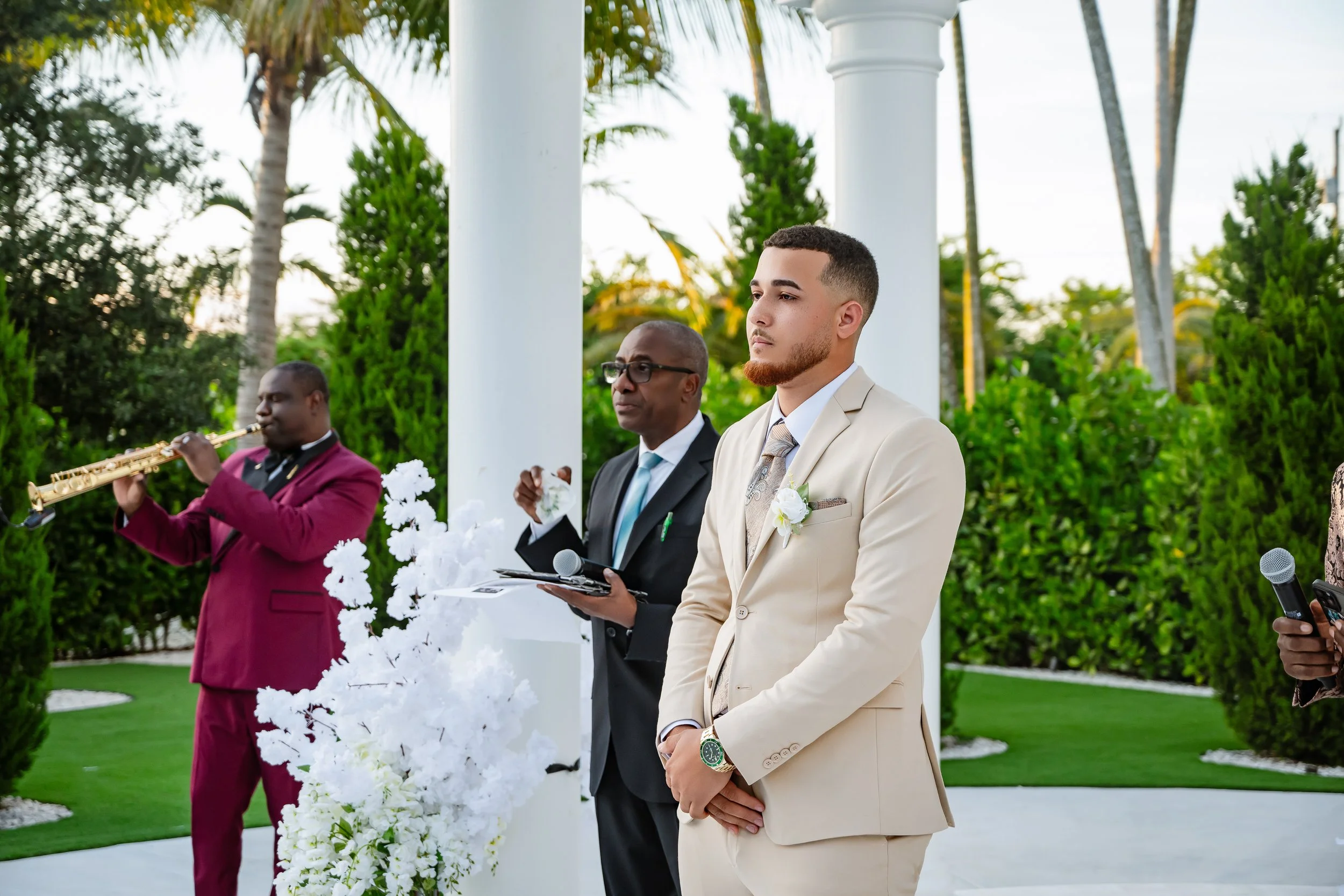 A groom in a cream suit with a boutonniere stands with hands clasped in front at an outdoor wedding ceremony, with a officiant, a musician, and a guest nearby in the background, surrounded by greenery and white columns.