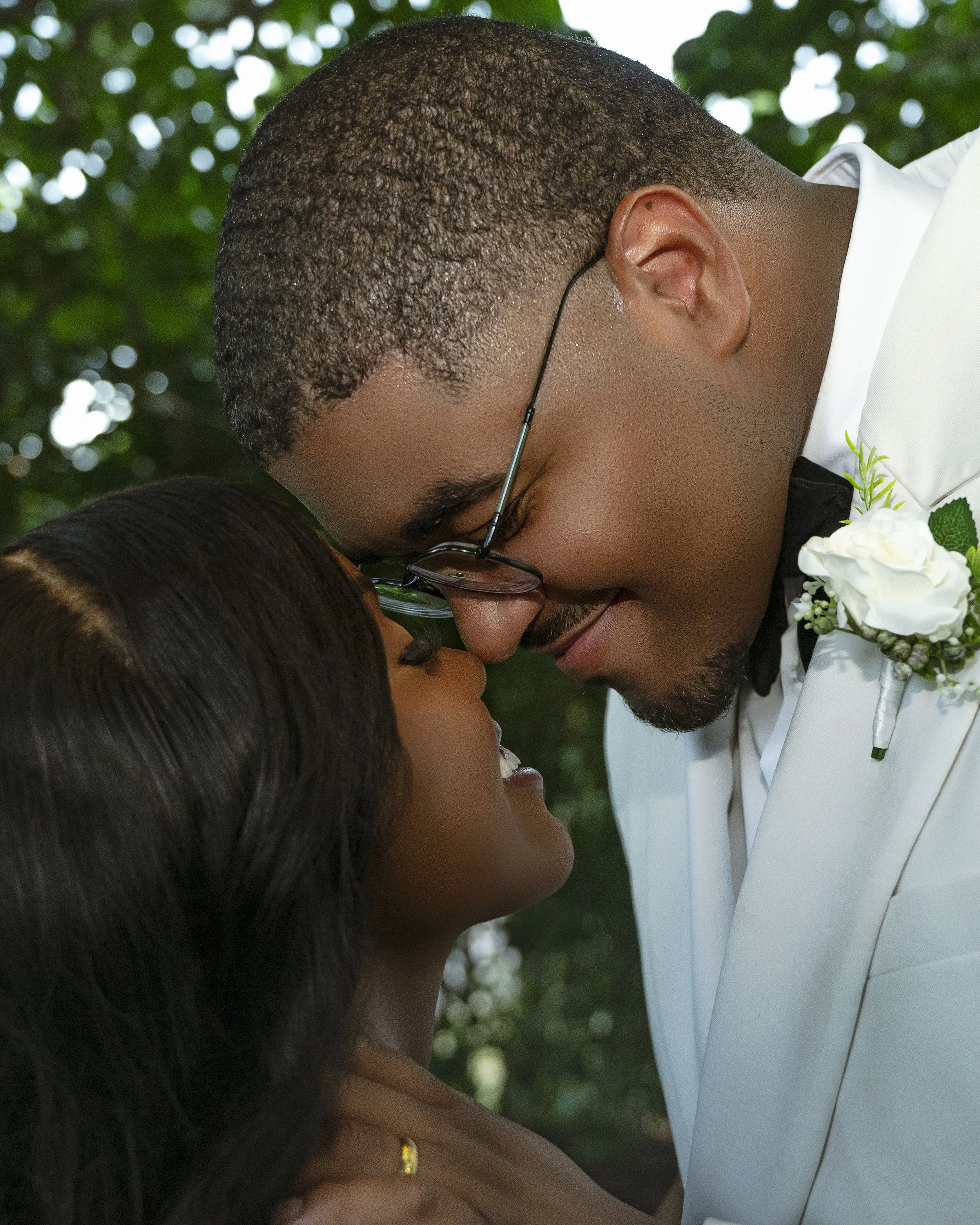 A close-up of a couple, a man and a woman, touching foreheads and smiling, outdoors with greenery in the background.