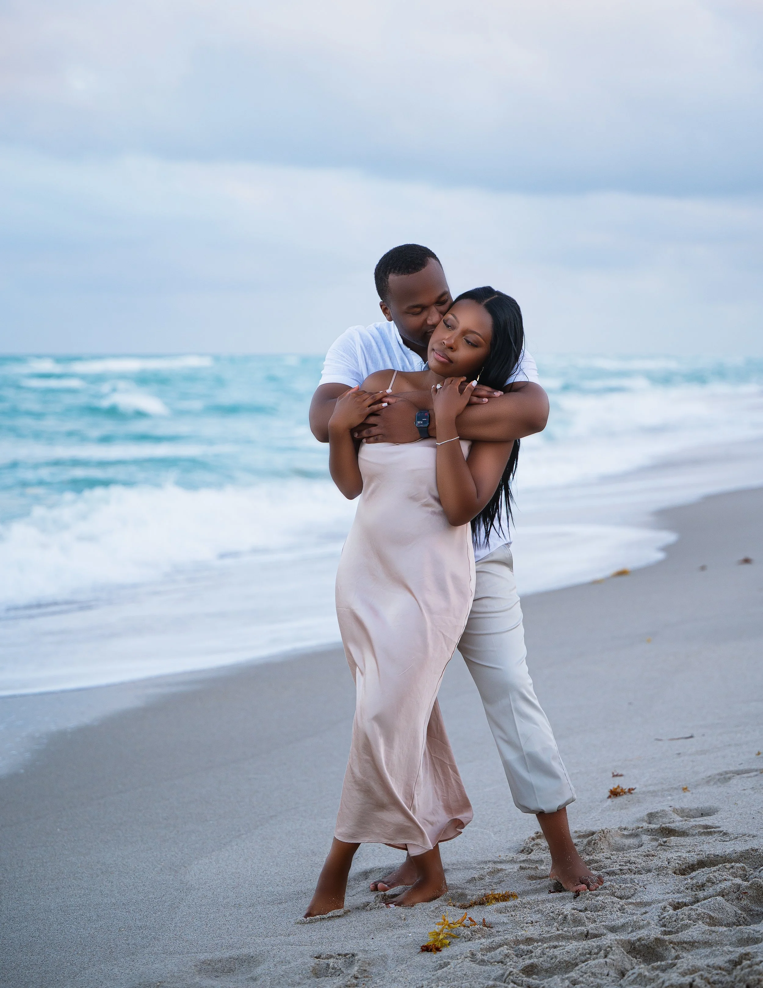 A loving couple embraces on the beach near the ocean, with the man standing behind the woman, both barefoot on the sand, under a cloudy sky.