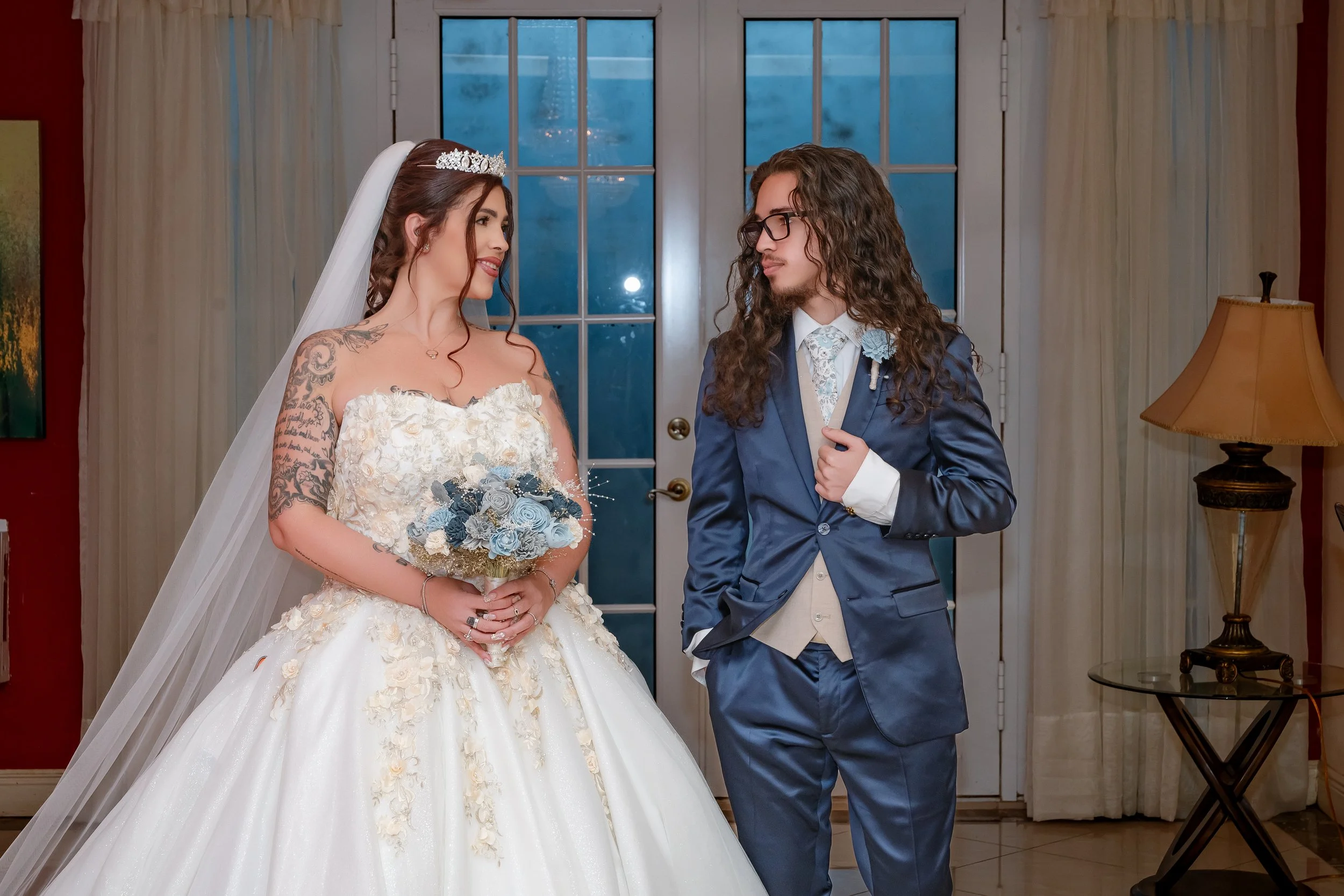 A bride in a white wedding gown and tiara holding a bouquet of blue and white flowers, standing next to a groom in a blue suit with glasses and long curly hair, inside a warmly decorated room.