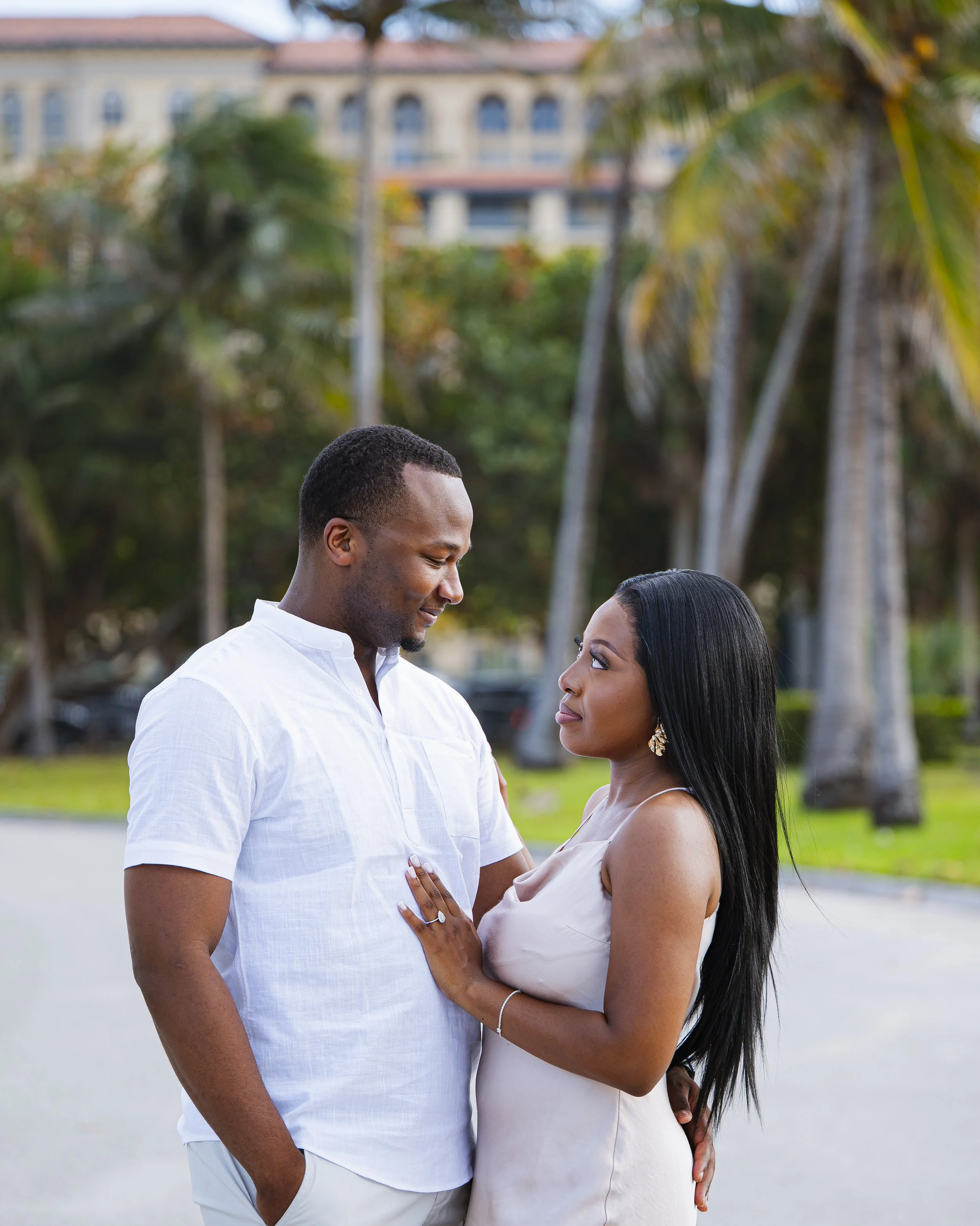 A romantic couple stands outdoors in a tropical setting with palm trees, gazing into each other's eyes.