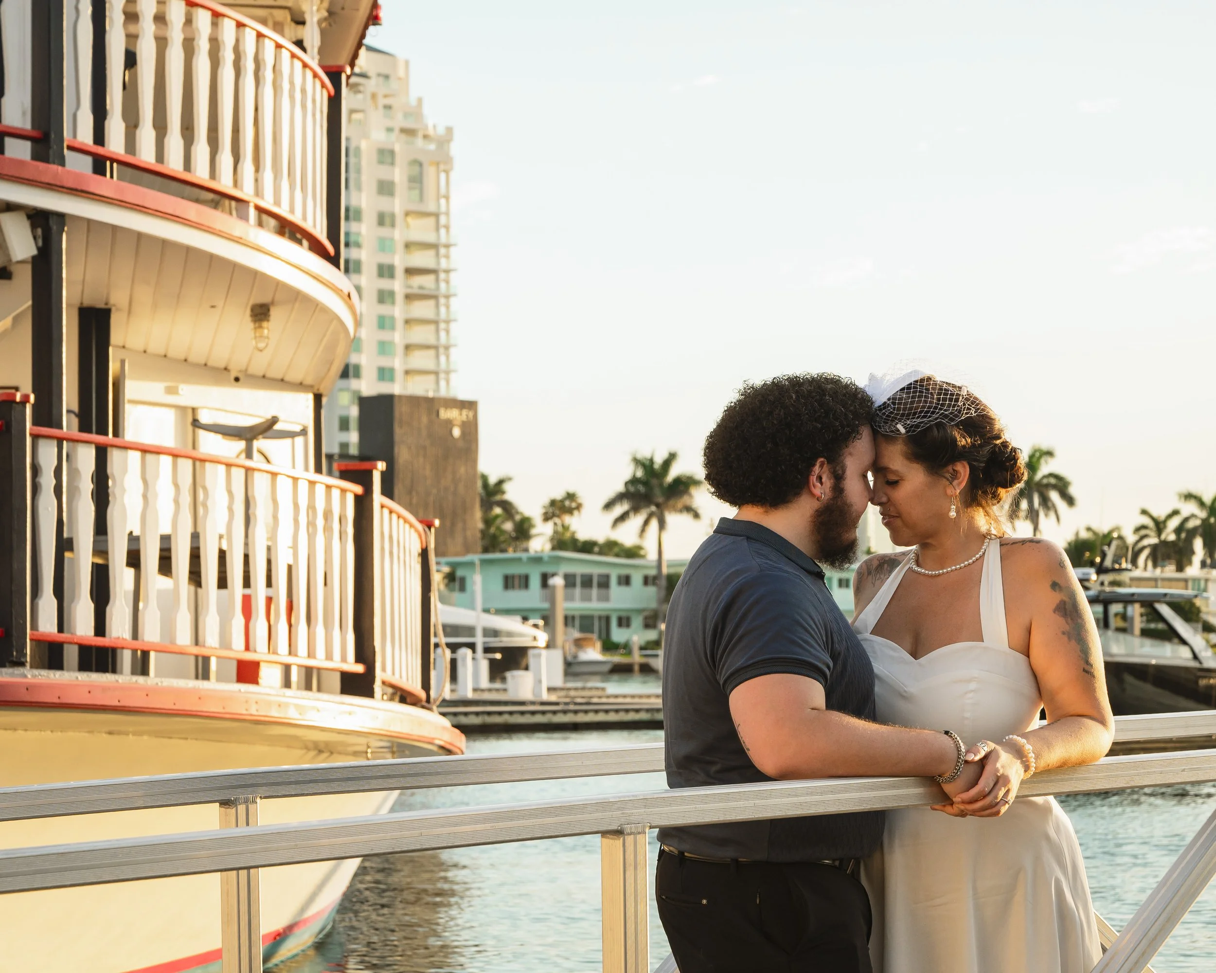 A couple with their foreheads touching on a boat dock with water and buildings in the background during sunset.