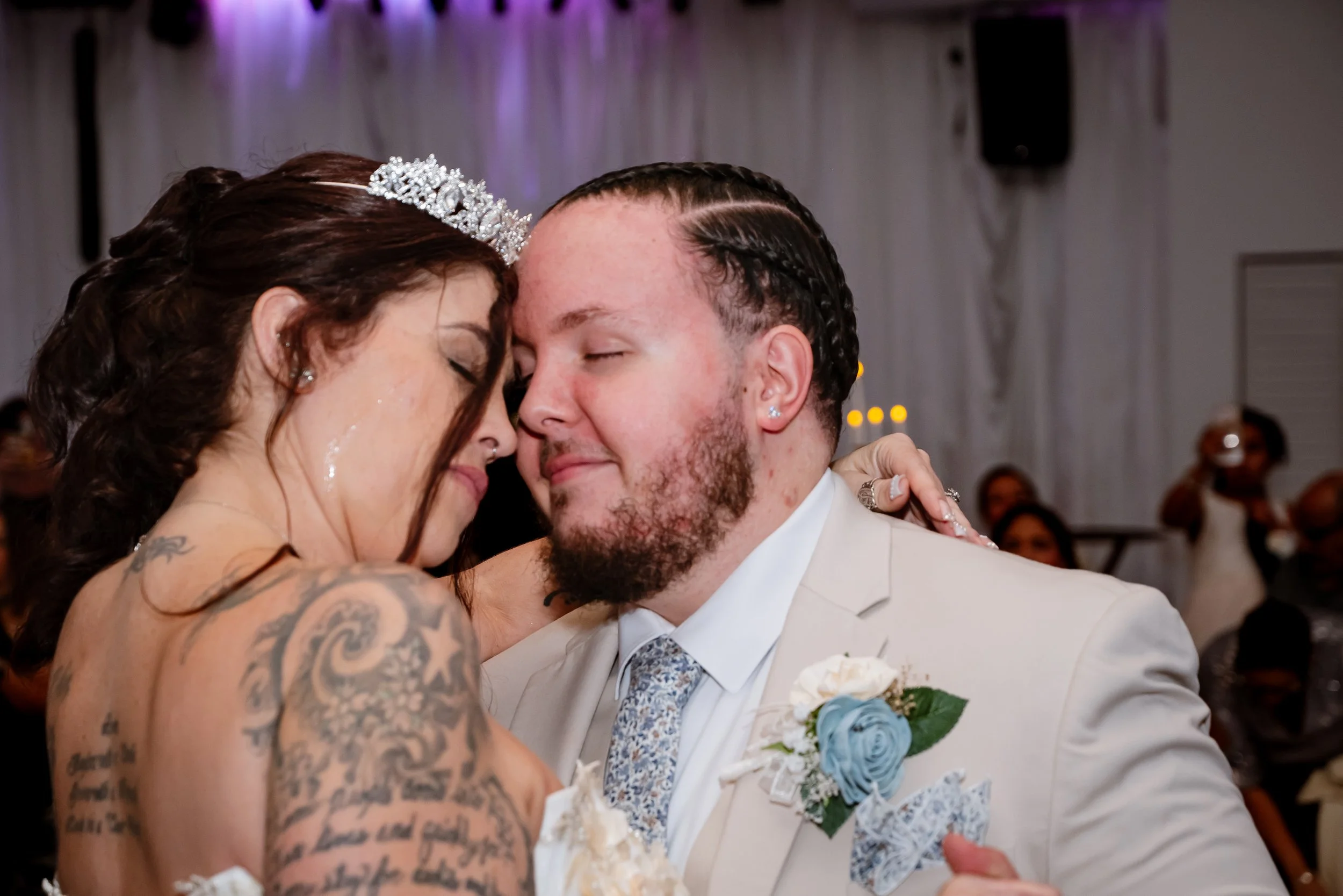 A bride and groom share a close, intimate moment at their wedding reception, with eyes closed and faces touching. The bride has brown hair, a tiara, and tattoos on her shoulders and arm, while the groom wears a light-colored suit, a patterned tie, an