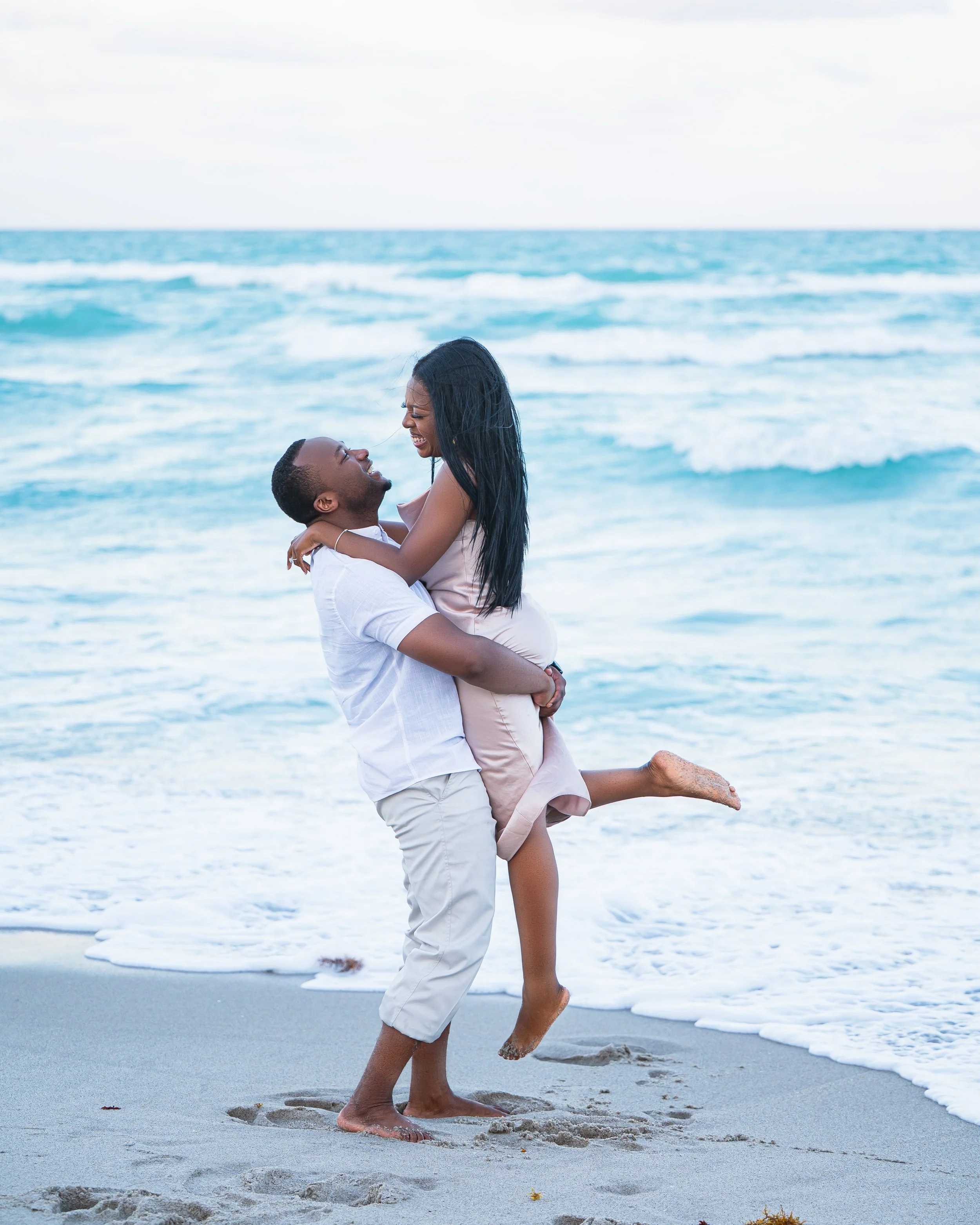 A happy couple on the beach with the man lifting the woman in his arms, both smiling, ocean waves in the background.