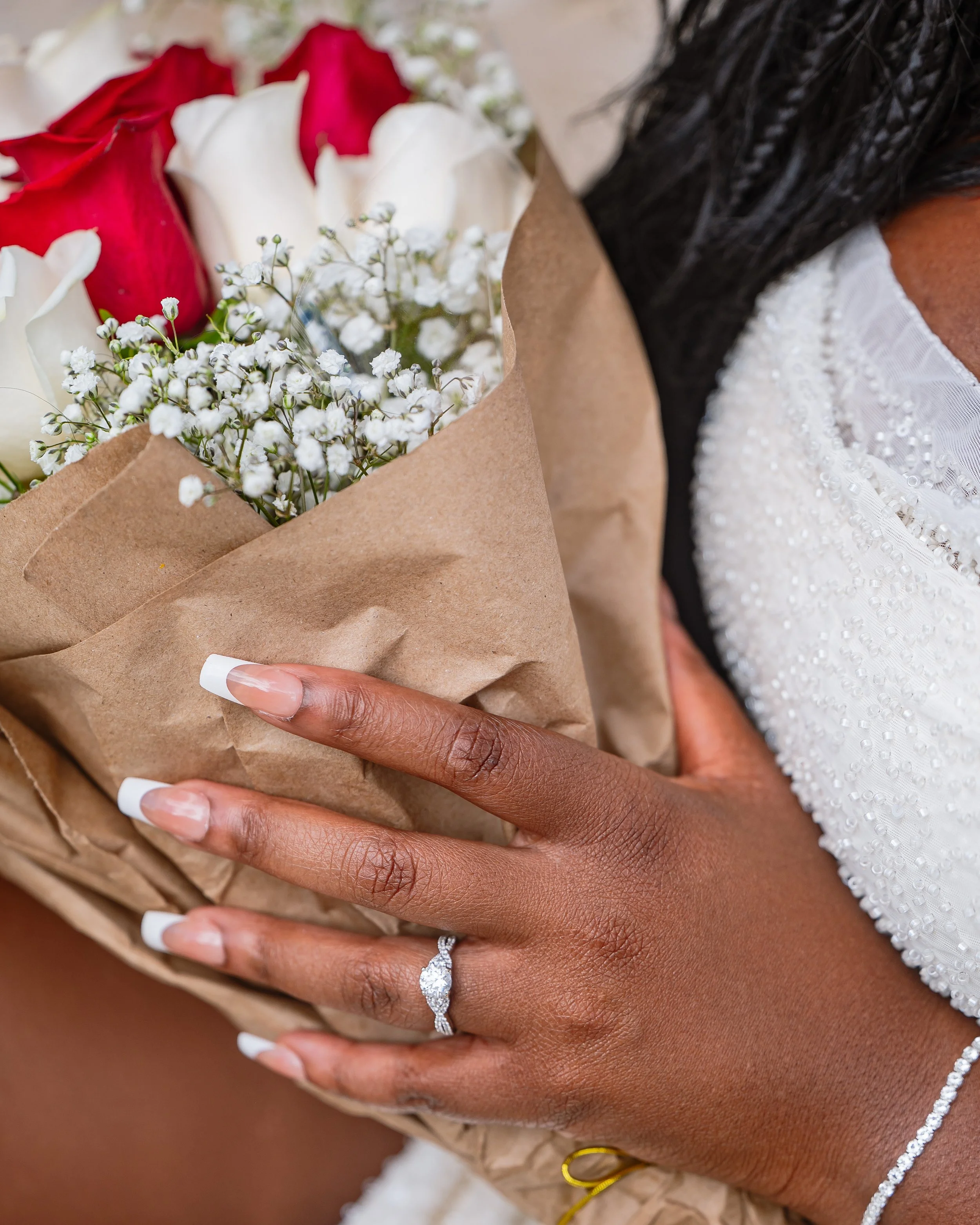 Person with a diamond ring on their finger holding a bouquet of red and white roses with white baby's breath, wrapped in brown paper.