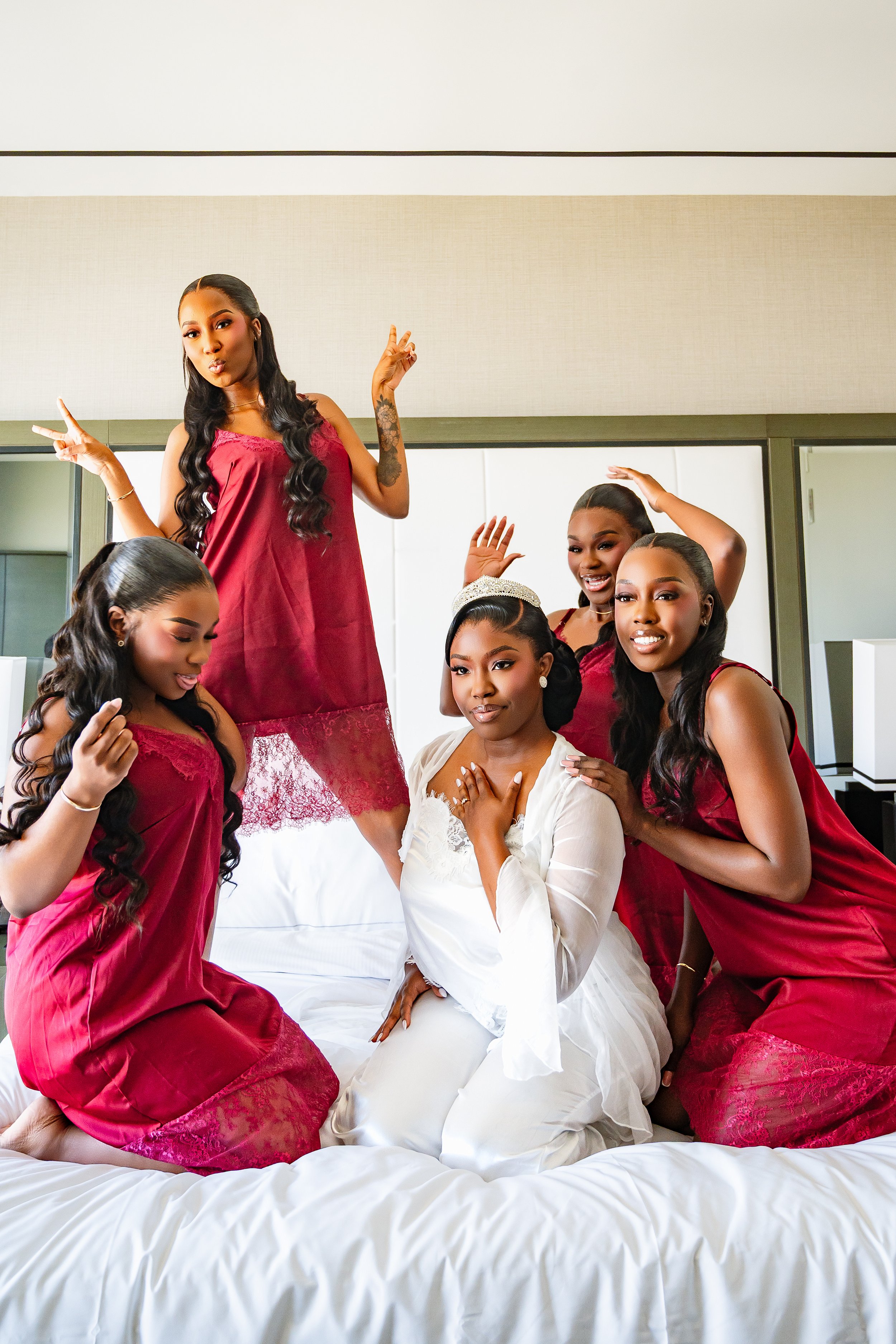 Group of women gathered in a bedroom, some wearing reddish dresses and one woman in white, with one woman in the center sitting on bed wearing white pajamas with a tiara, surrounded by others in matching dresses, all smiling and posing.
