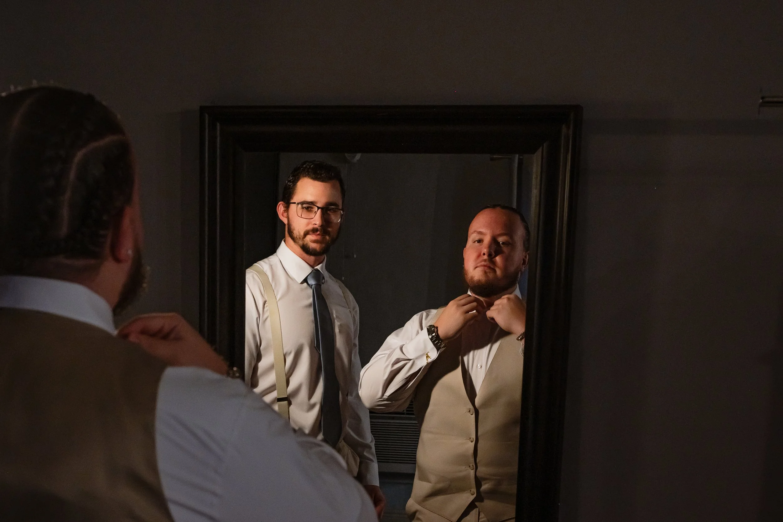 Two men looking at their reflection in a mirror, one adjusting his collar, in a dimly lit room.