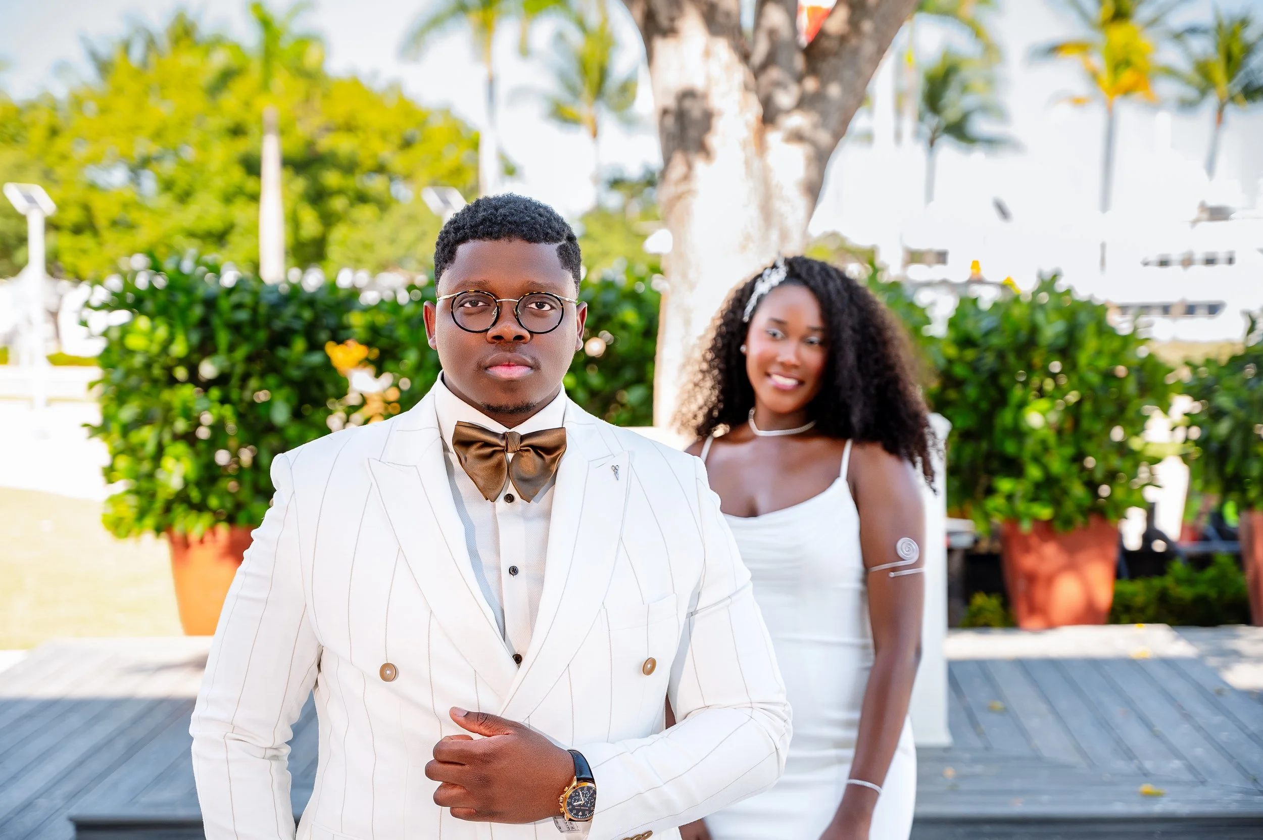 A young man in a white suit with thin stripes, glasses, and a bow tie posing for the camera, with a young woman in a white dress standing behind him smiling outdoors near a tree and large potted plants.