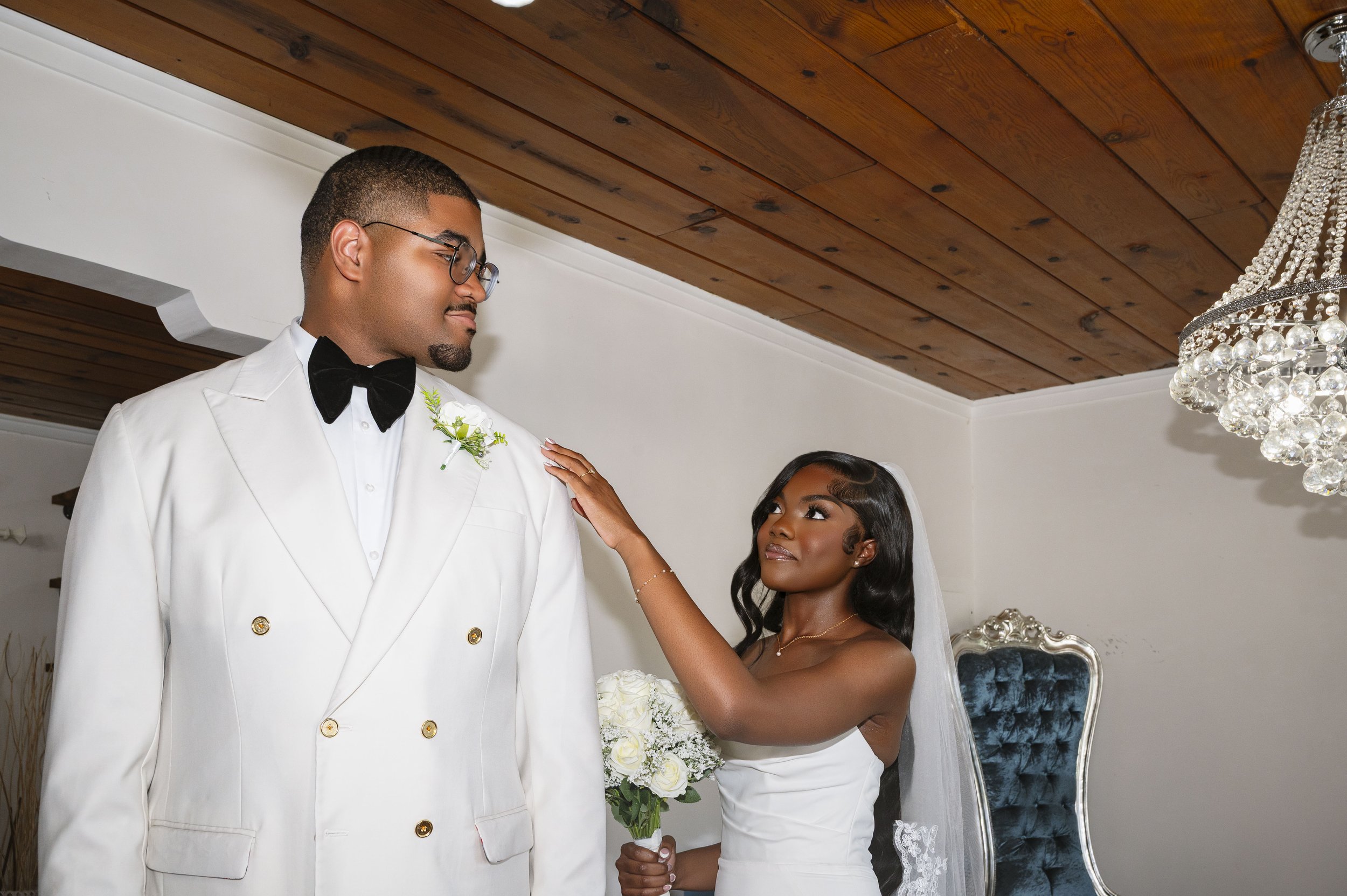 Bride touching groom's shoulder during wedding ceremony indoors, with chandelier and wooden ceiling in background.