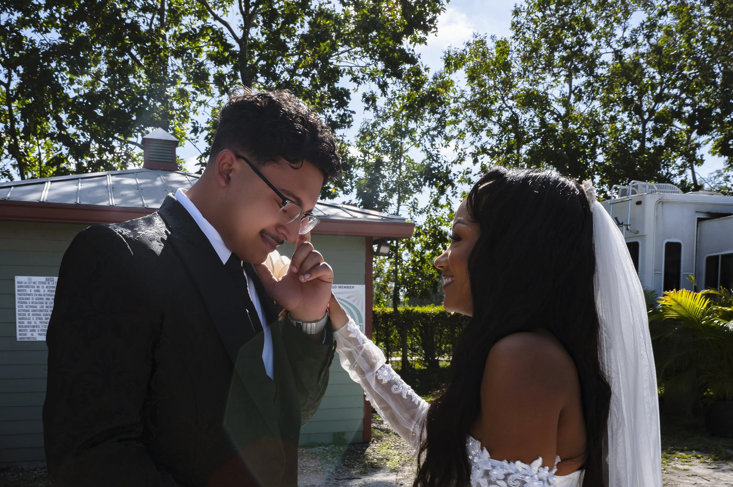 A bride and groom smiling at each other outdoors, with trees and a trailer in the background. The groom is wearing a black suit and glasses, and the bride has long dark hair and a white wedding dress with a veil.