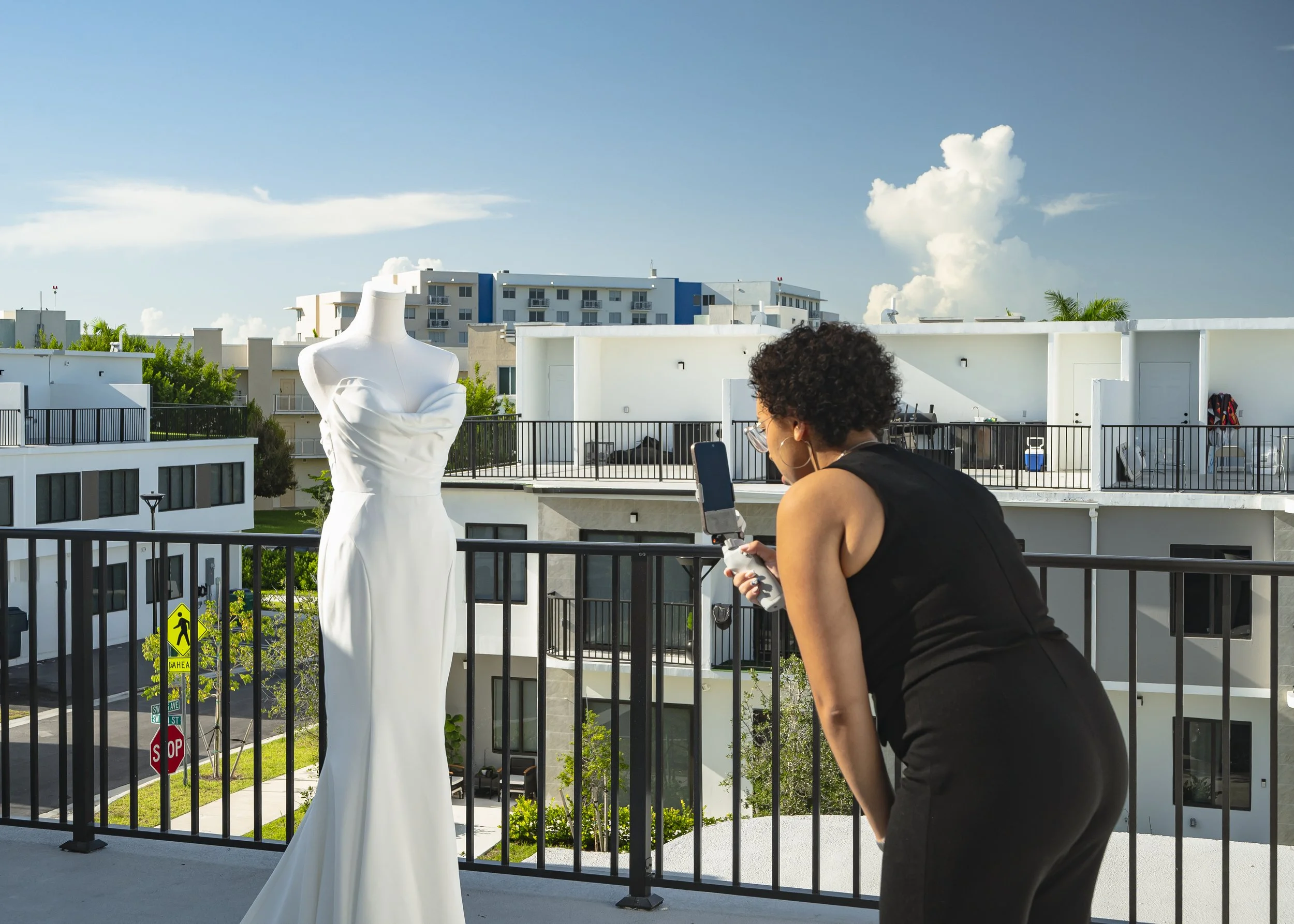 A woman with curly hair and glasses takes a photo of a white dress on a mannequin on a balcony.