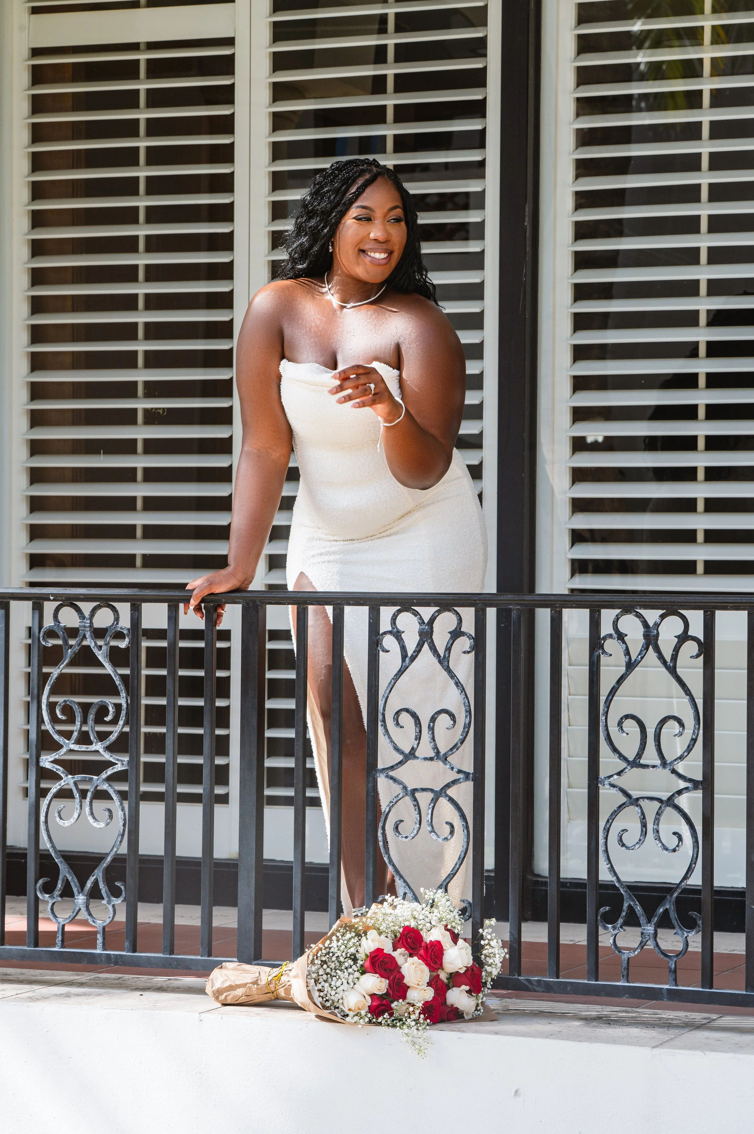 Woman in white dress smiling outdoors on a balcony with a bouquet of red and white roses on the ground.
