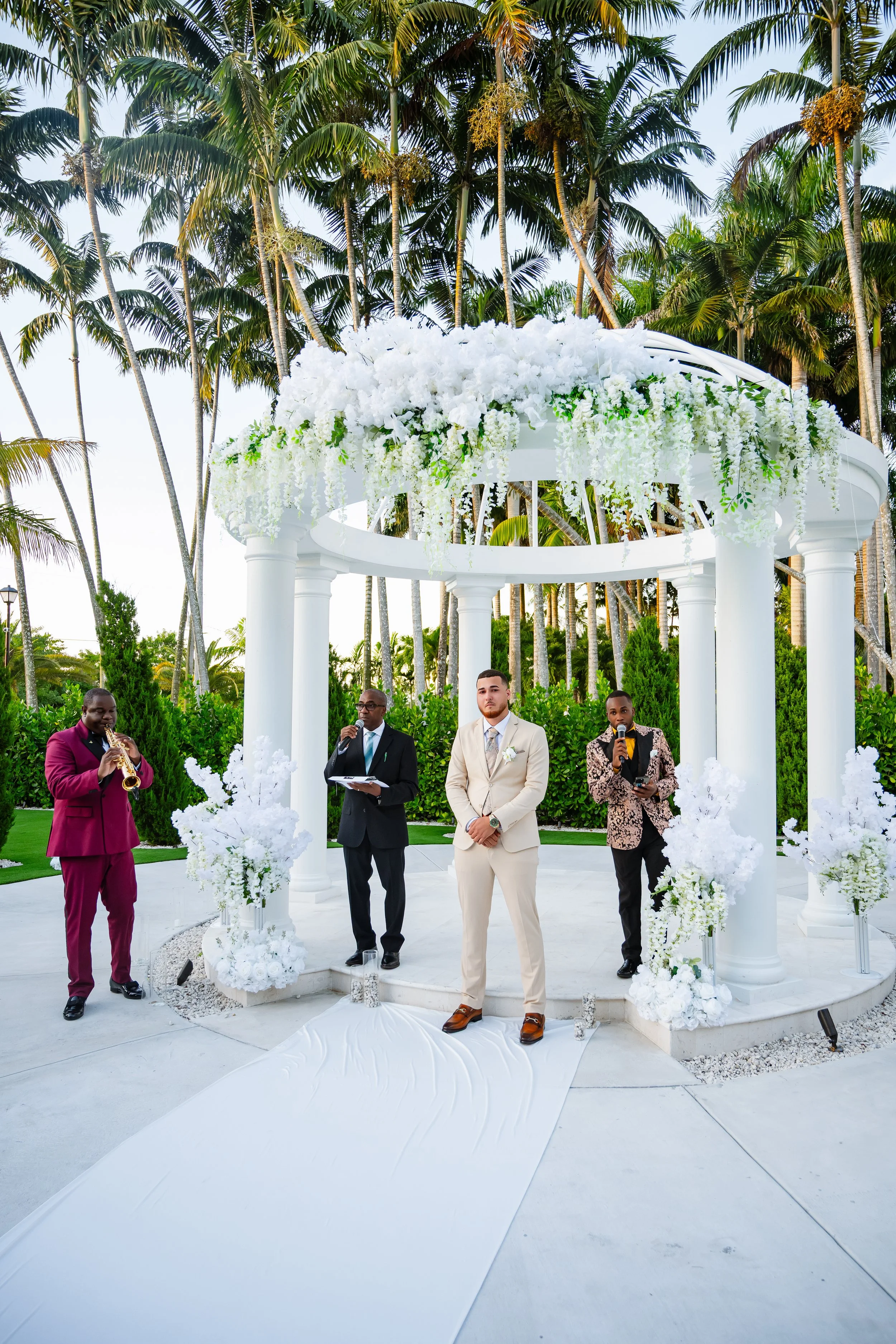 A wedding ceremony taking place outdoors under a white floral gazebo with four white columns, surrounded by tall palm trees and lush green bushes. Four men are standing in front of the gazebo: one in a beige suit, another in a black suit holding a mi