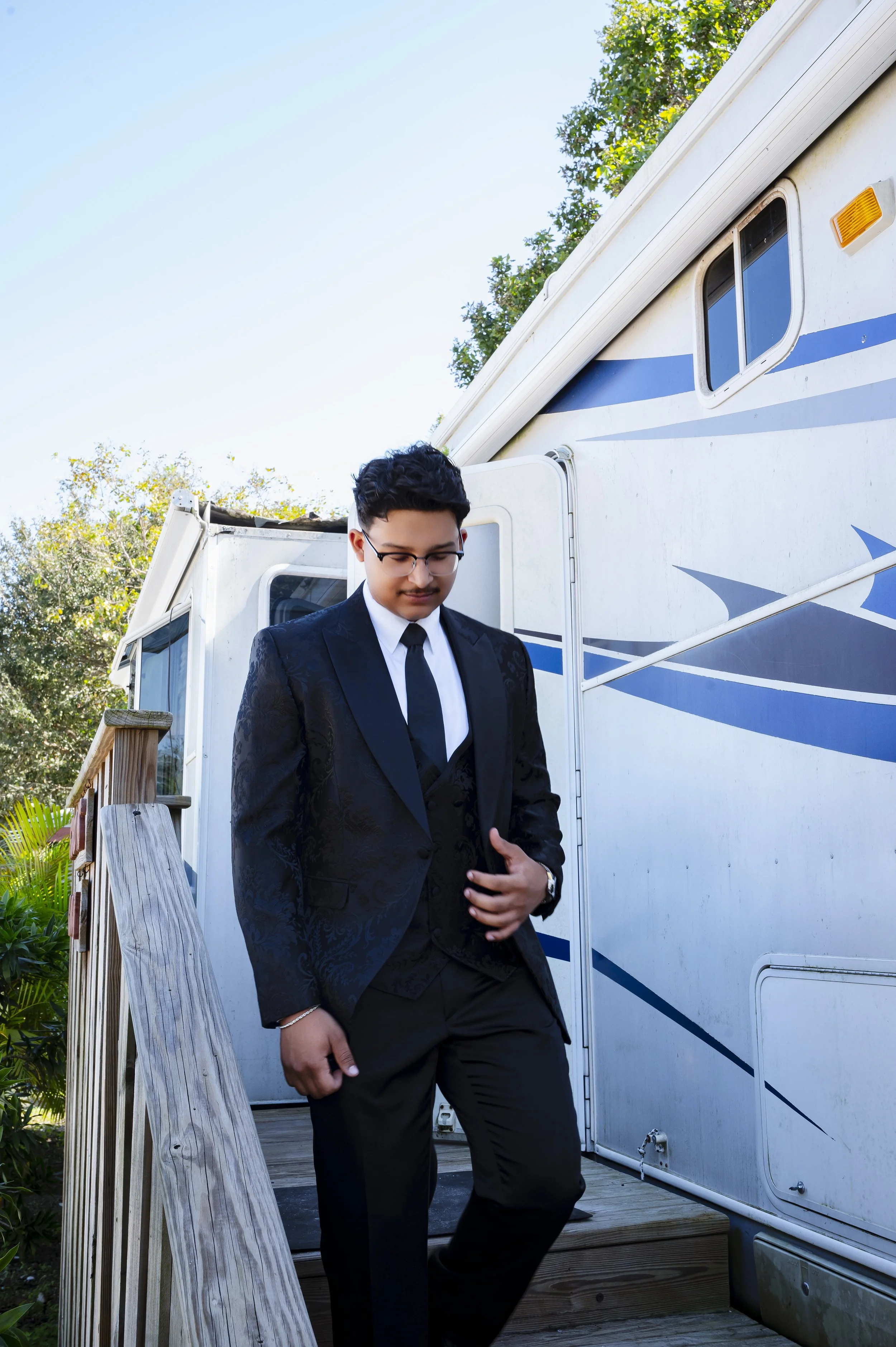 A young man dressed in a black suit with a white shirt and black tie, wearing glasses, standing outside a white camper trailer with blue stripes, on a wooden deck surrounded by greenery.