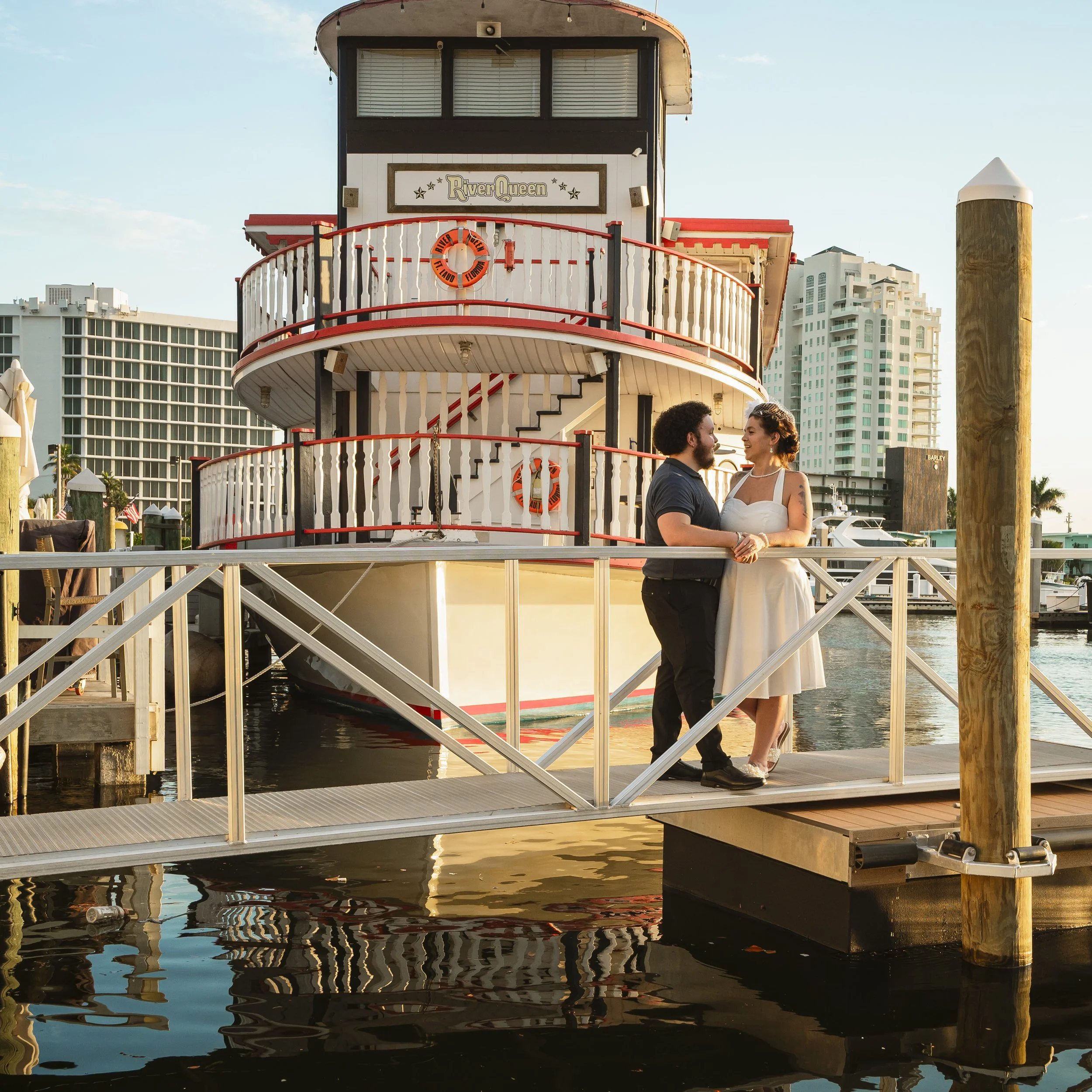 A couple stands on a dock in front of a riverboat named River Queen, with modern high-rise buildings in the background during sunset.